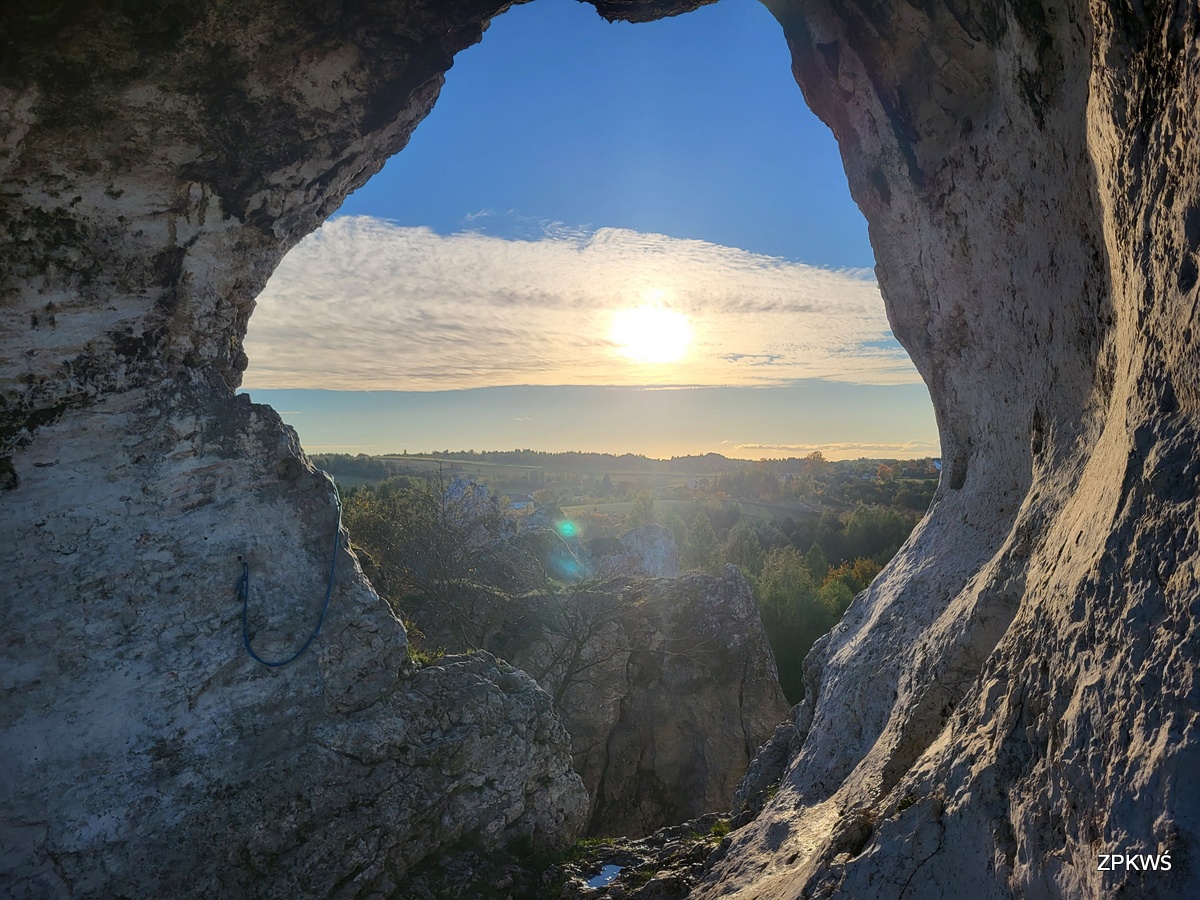 Fotografia przedstawia widok na krajobraz jurajski widoczny przez okno skalne. Zdjęcie zrobione podczas pięknego słonecznego dnia. W tle widoczne jurajskie wychodnie skalne, łąki i lasy. 