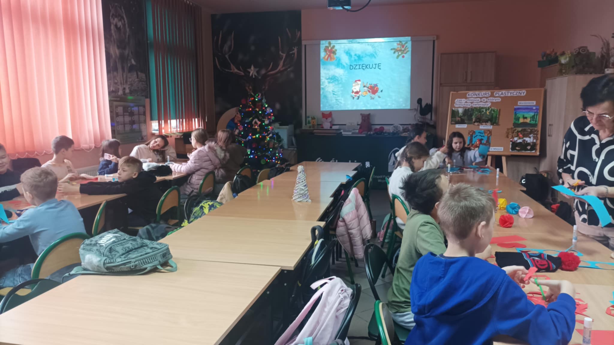 Dzieci siedzą przy stolikach w Ośrodku Edukacyjnym i wykonują bombki z papieru, w tle prezentacja o tematyce świątecznej oraz choinka.