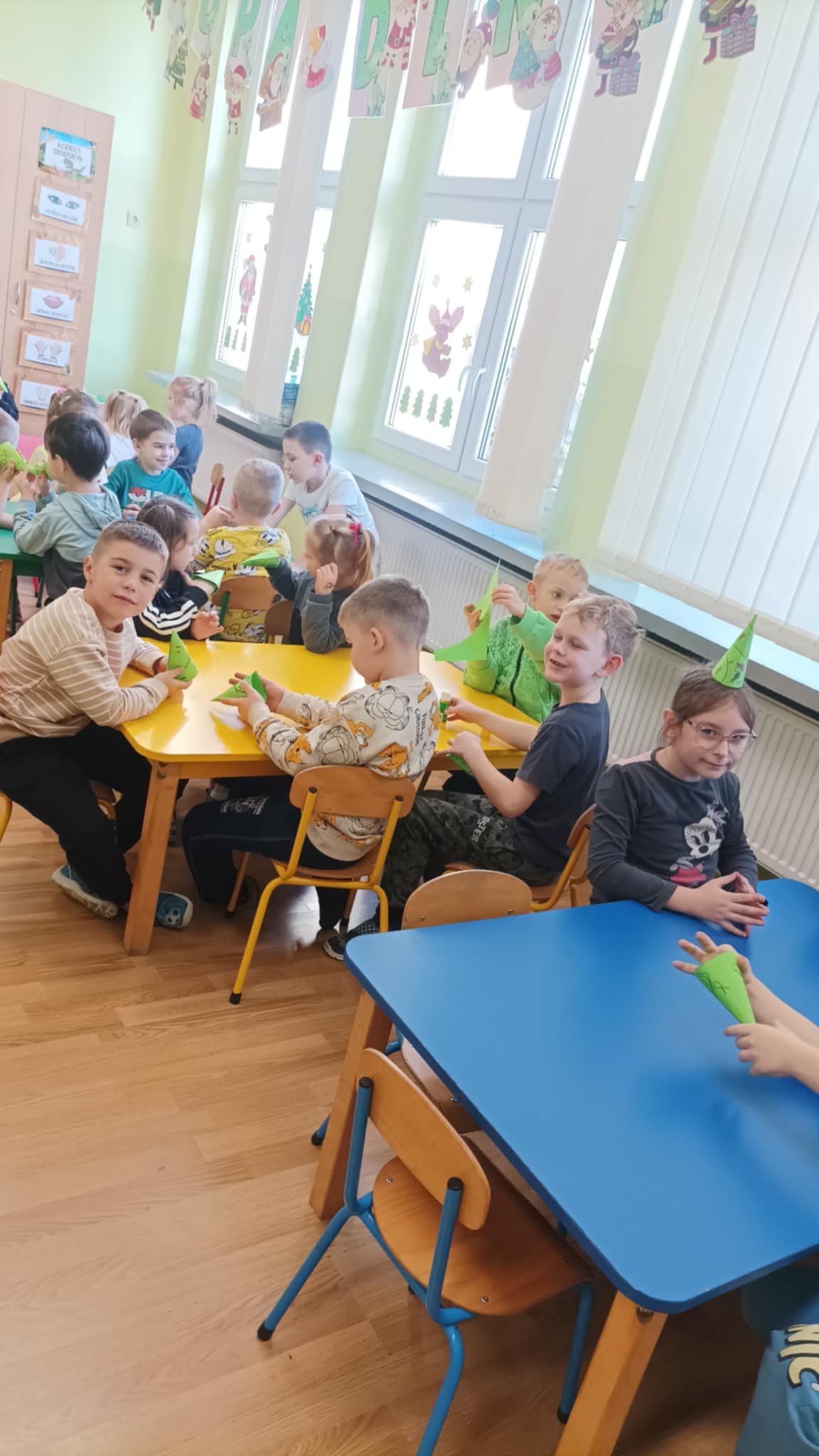 Przedszkolaki siedzą przy stolikach i pokazują gotową choinkę z papieru.