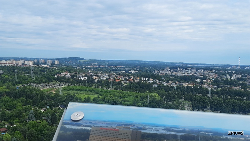 Zdjęcie zrobione jest z tarasu widokowego, które przedstawia panoramę miasta. Na pierwszym planie widać część tablicy ze zdjęciem miasta. Na drugim planie widać drzewa, domy, bloki, słupy eklektyczne i kominy z zakładu hutniczego. Niebo jest pochmurne niebiesko białe.  