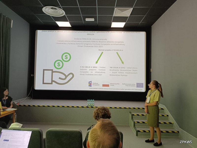 Na zdjęciu widoczna jest sala konferencyjna podczas prezentacji projektu „Dobre praktyki w zakresie ochrony cennych przyrodniczo siedlisk i gatunków w PK ‘Orlich Gniazd’”. Na ekranie wyświetlony jest slajd z informacjami o finansowaniu projektu: całkowita wartość to 4 720 806 zł, z czego 85% (4 012 685,10 zł) pochodzi z Funduszu Spójności programu Fundusze Europejskie na Infrastrukturę, Klimat i Środowisko 2021–2027, a 15% (708 120,90 zł) stanowi wkład własny beneficjenta – Województwa Śląskiego / Zespołu Parków Krajobrazowych Województwa Śląskiego. Po prawej stronie kadru stoi prelegentka w zielonym stroju, przedstawiająca dane dotyczące finansowania, natomiast po lewej przy stole siedzi osoba obsługująca laptop. Na pierwszym planie widać uczestników konferencji.