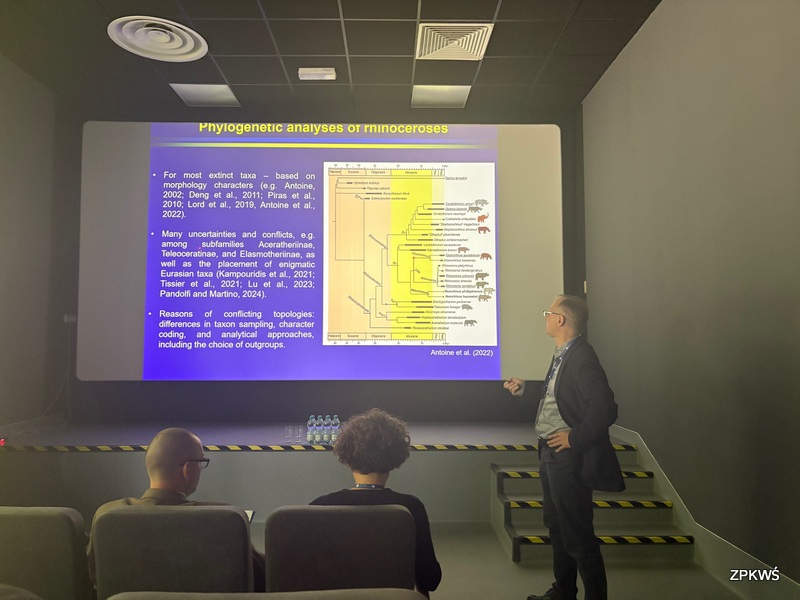 Na zdjęciu widoczna jest sala konferencyjna w trakcie wykładu naukowego. Prelegent – stojący po prawej stronie w garniturze – prezentuje slajd zatytułowany „Phylogenetic analyses of rhinoceroses”. Na ekranie znajduje się schemat drzewa filogenetycznego nosorożców oraz punkty opisujące niepewności i konflikty w analizach taksonomicznych. Na pierwszym planie widać uczestników konferencji siedzących w fotelach, słuchających 