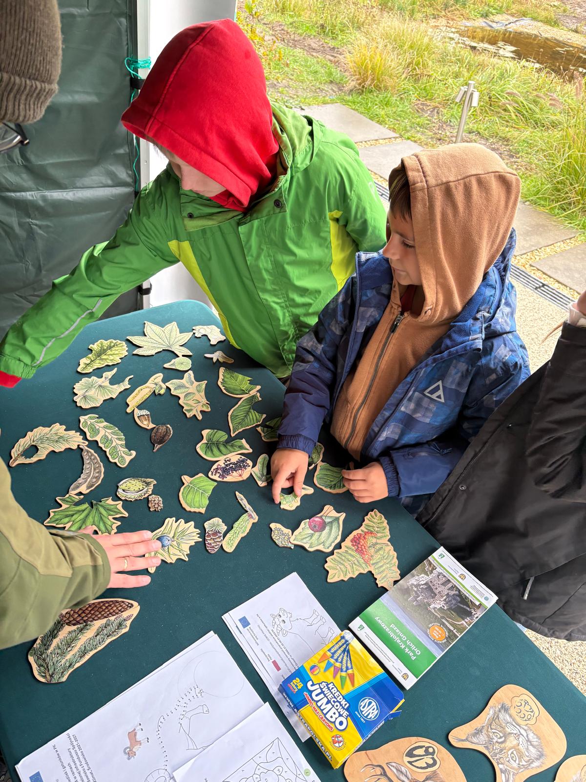 Na zdjęciu widać dwójkę dzieci ubraną w ciepłe kurtki z kapturami — jedno ma czerwoną czapkę pod zieloną kurtką, drugie ma brązową bluzę z kapturem i niebieską kurtkę. Stoją przy stole przykrytym ciemnozielonym obrusem. Na stole rozłożone są drewniane elementy w kształcie różnych liści, szyszek i owoców leśnych, wyglądające jak elementy układanki edukacyjnej. Widać również kredki Jumbo, kolorowanki oraz broszurę informacyjną dotyczącą przyrody. Dzieci zdają się aktywnie uczestniczyć w zabawie edukacyjnej na świeżym powietrzu. W tle widać fragment ścieżki i trawy.