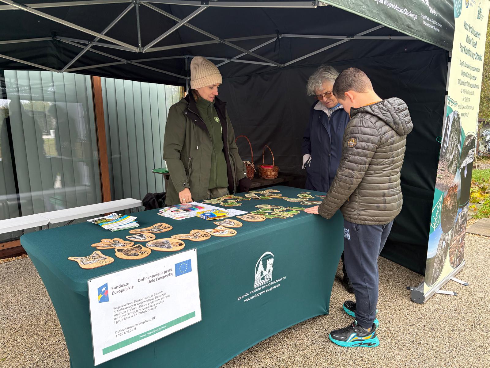 Na zdjęciu widać stoisko edukacyjne ustawione pod zielonym namiotem plenerowym. Za stołem stoją dwie osoby — kobieta w zielonej kurtce i beżowej czapce oraz starsza osoba w ciemnej kurtce. Po drugiej stronie stołu stoi chłopiec ubrany w ciemną pikowaną kurtkę, granatowe spodnie i sportowe buty. Na stole leży ciemnozielony obrus z logo Zespołu Parków Krajobrazowych Województwa Śląskiego. Rozłożone są drewniane elementy przedstawiające liście, owoce i inne motywy przyrodnicze, a także kredki Jumbo i materiały edukacyjne. Na froncie stołu widnieje tabliczka z informacją o dofinansowaniu projektu przez Unię Europejską (Fundusze Europejskie). W tle widać baner informacyjny oraz elementy architektury i zieleni.