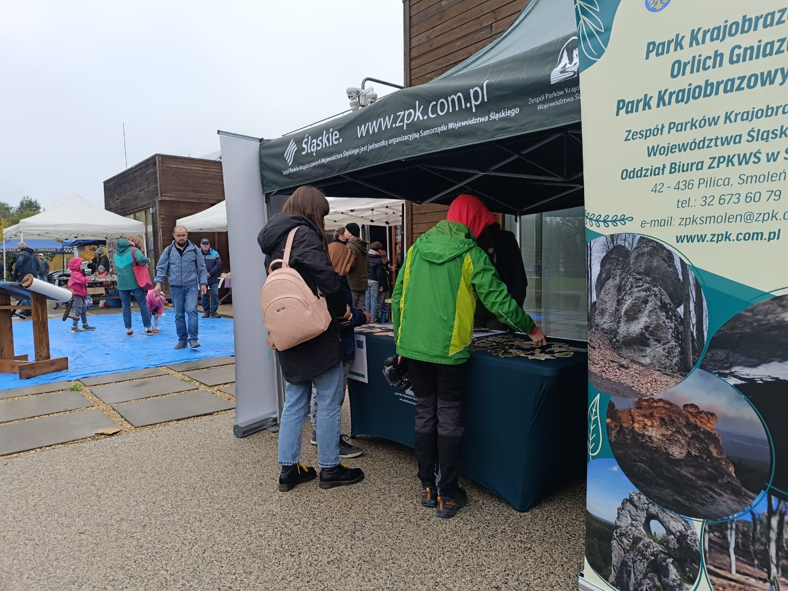 Na zdjęciu widać dziecko w kolorowej kurtce, które stoi przy stole z ciemnozielonym obrusem. Obok niego znajduje się osoba dorosła w ciemnej kurtce z jasnym plecakiem , pochylająca się nad stołem. Na stole rozłożone są drewniane elementy przedstawiające liście, owoce i szyszki, a także kredki Jumbo i kartki do kolorowania. Dziecko i dorosły wspólnie układają lub dopasowują drewniane elementy, uczestnicząc w aktywności edukacyjnej na świeżym powietrzu. 