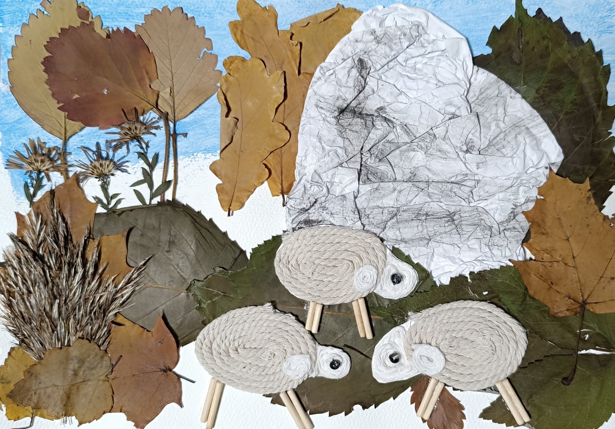 Na zdjęciu znajduje się praca plastyczna przedstawiająca trzy owce na tle jesiennego krajobrazu. Owce wykonane są z białego sznurka uformowanego w spiralę, z głowami z mniejszych zwojów i czarnymi oczkami, a ich nogi zrobione są z drewnianych patyczków. Tło składa się z suszonych liści w różnych odcieniach brązu, zieleni i żółci, które tworzą drzewa, krzewy i trawę. W prawym górnym rogu znajduje się duży biały element przypominający skałę wapienną wykonaną z pogniecionej kartki papieru. Na górze pracy widać niebieskie niebo, a całość ma charakter naturalny i jesienny, z wykorzystaniem materiałów z przyrody.