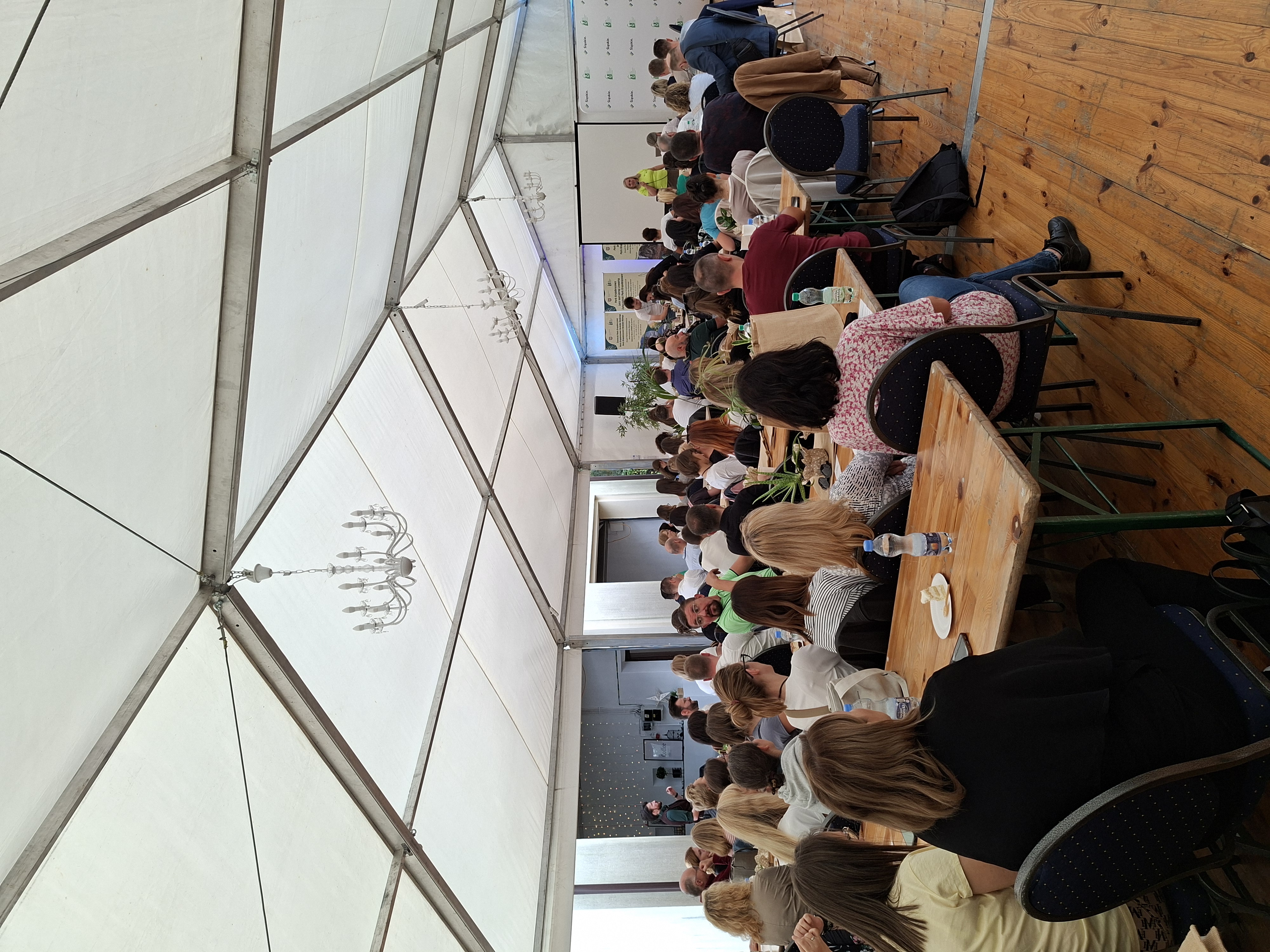 Na zdjęciu widoczne wnętrze białej hali namiotowej, w której przy drewnianych stołach siedzą ludzie (ok. 80 osób) patrzący na kobietę w jasnozielonej koszulce. Kobieta stoi przed siedzącymi i trzyma mikrofon (wita uczestników spotkania).