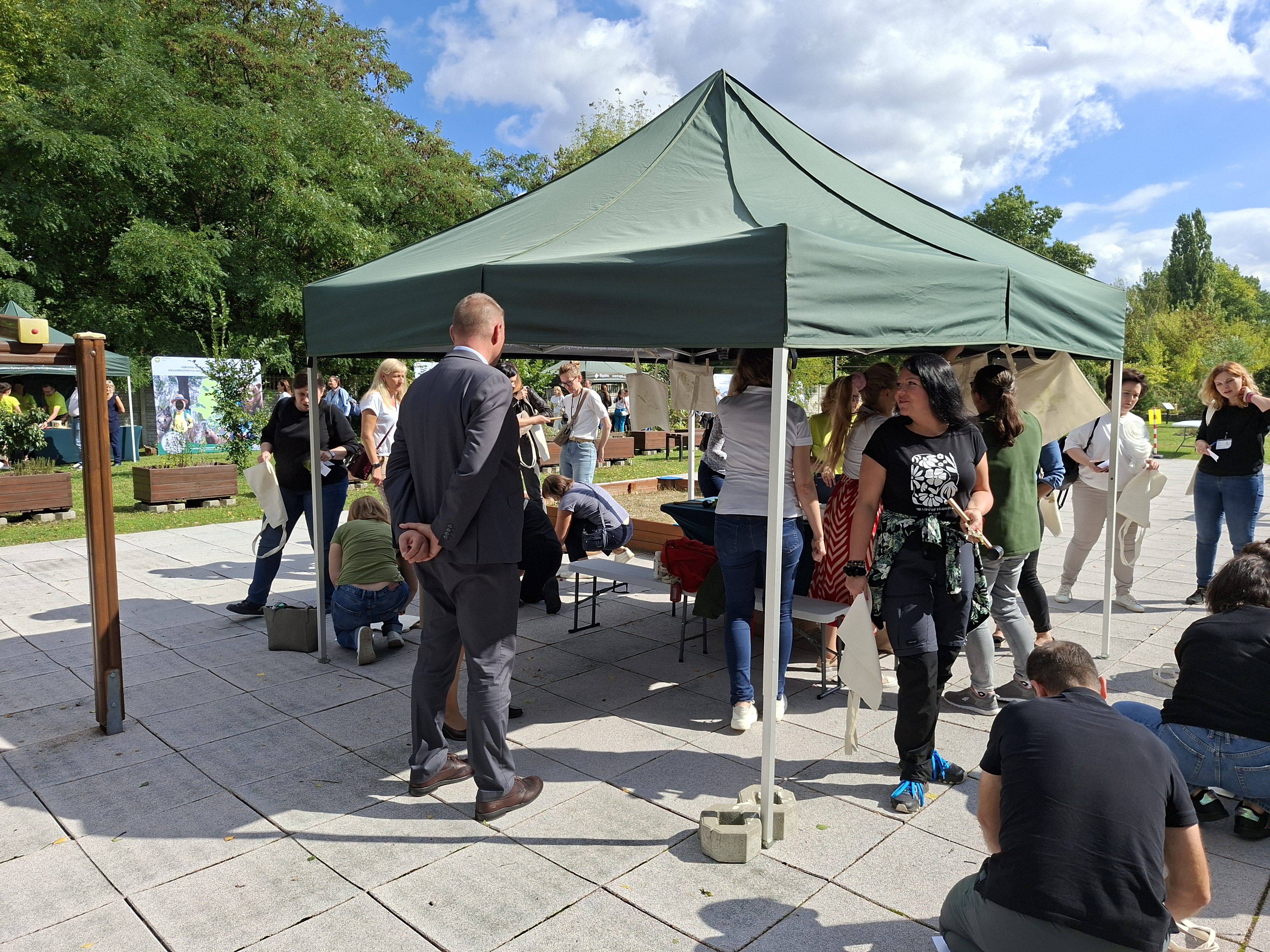 Na pierwszym planie widoczny mężczyzna w ciemnoszarym garniturze stojący tyłem do fotografa. W tle zielony namiot, pod dachem którego grupa osób wykonuje zadania manualne - odbijają zielone liście na płóciennych torbach, przy pomocy gumowych młotków.