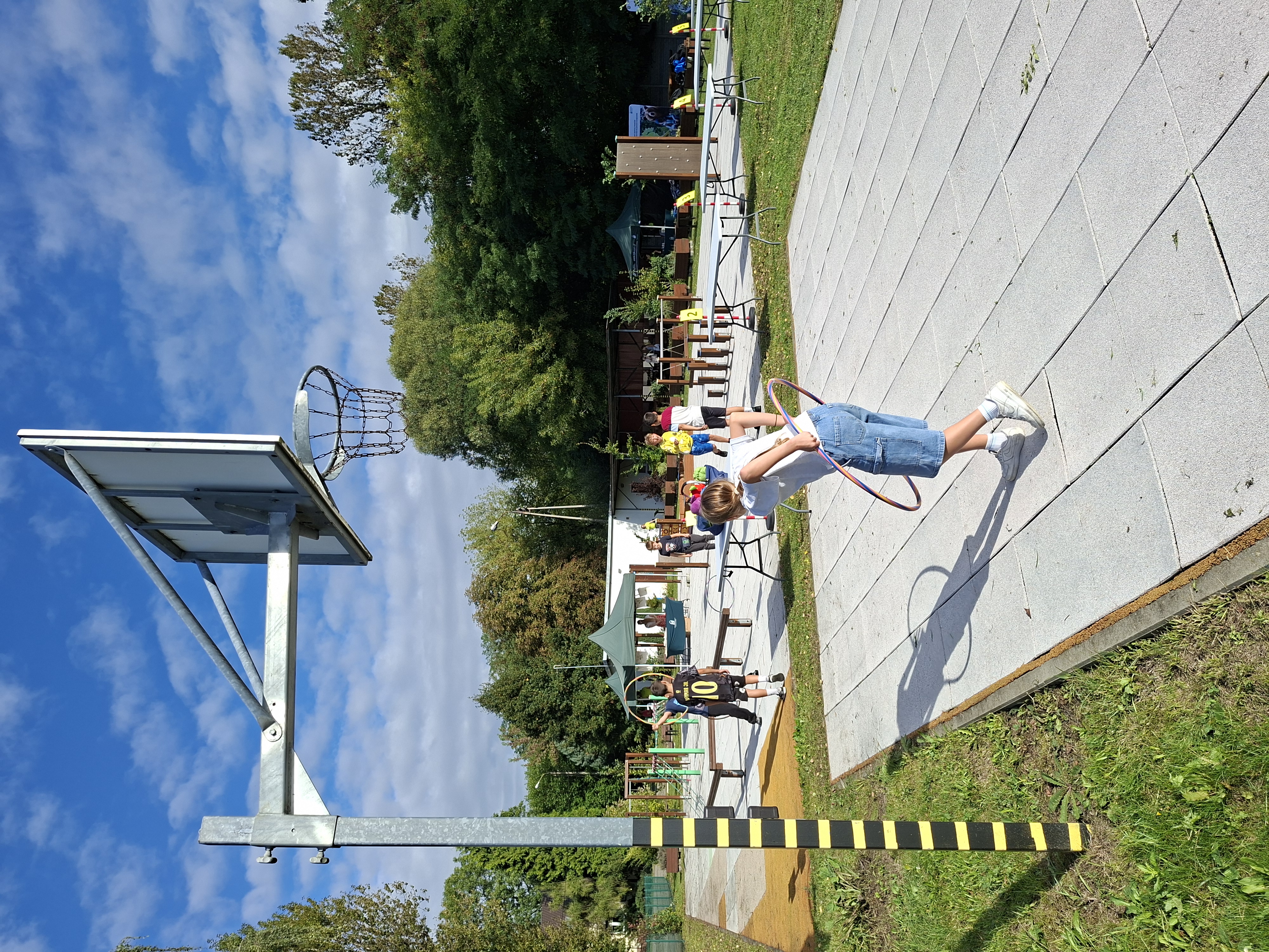 Na zdjęciu widoczny kosz do gry w koszykówkę. Pod koszem w pochylonej pozycji stoi chłopiec ubrany w jasną koszulkę i dżinsowe spodenki, który bawi się hula-hop. W tle widoczny plac zabaw, na którym bawi się młodzież. Plac otoczony zielonymi drzewami.