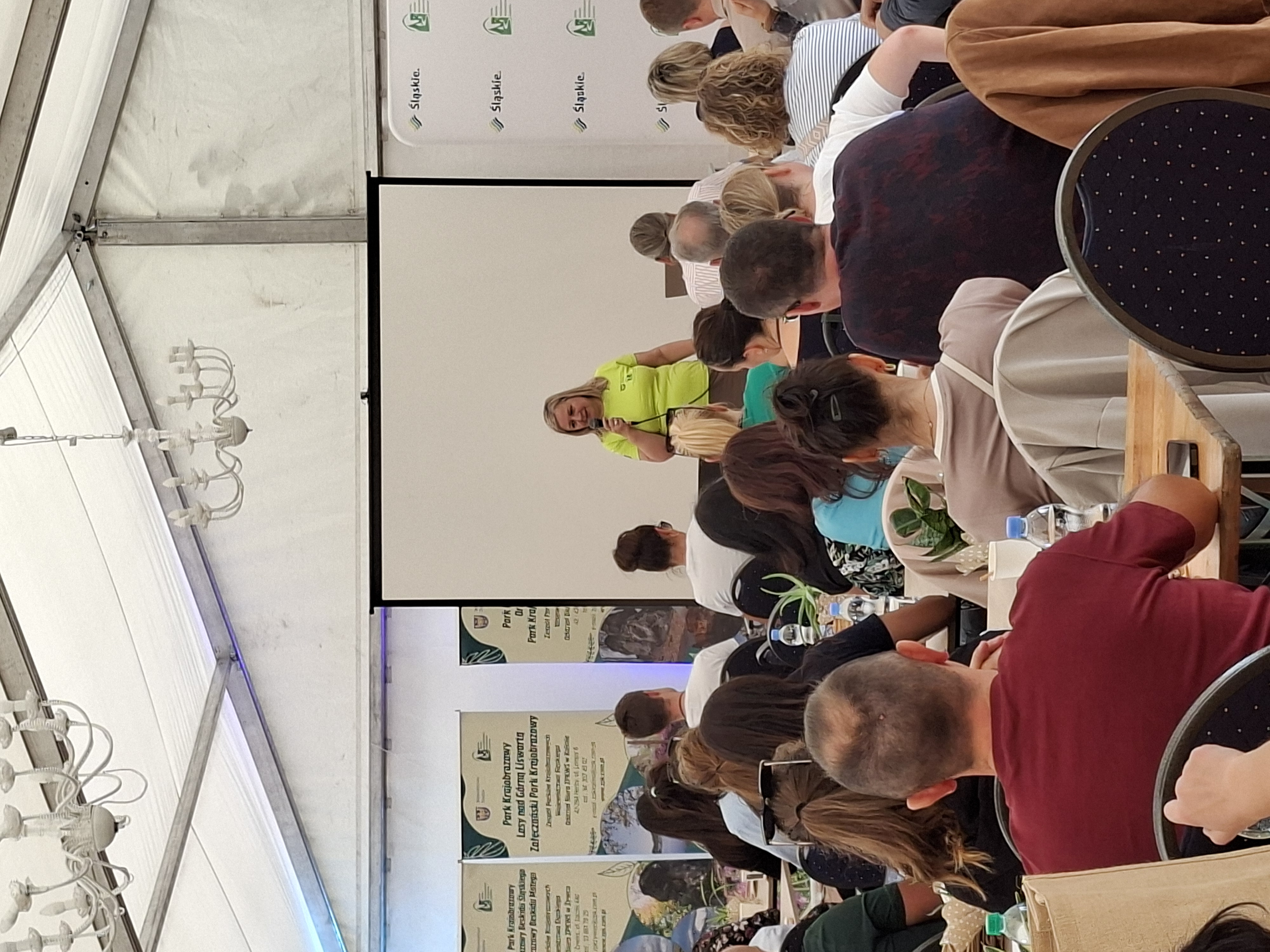 Na zdjęciu, na pierwszym planie widać od tyłu siedzące na krzesłach osoby. Na drugim planie, przed siedzącymi osobami, stoi kobieta w jasnozielonej koszulce, trzymająca mikrofon. Kobieta stoi na tle ekranu do wyświetlania prezentacji multimedialnych.