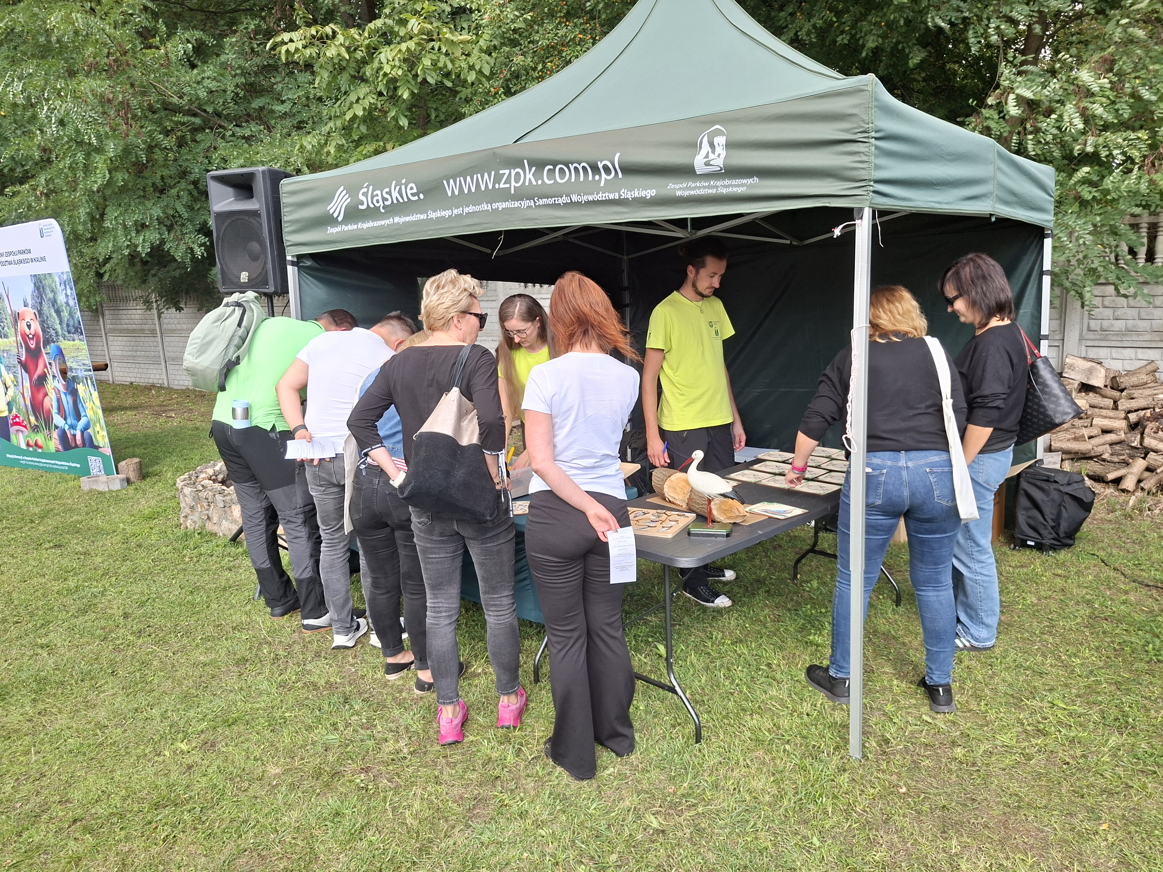 Na zdjęciu widoczny zielony namiot, pod którym stoją stoły. Na stołach leżą pomoce dydaktyczne - gry, puzzle i łamigłówki. Przed namiotem stoi grupa osób (kobiety i mężczyźni), który zajęci są rozwiązywaniem zadań, w oparciu o pomoce dydaktyczne leżące na stołach. Pod namiotem widoczne osoby - mężczyzna i kobieta - w jasnozielonych koszulkach, którzy tłumaczą zgromadzonym zasady rozwiązywania zadań.