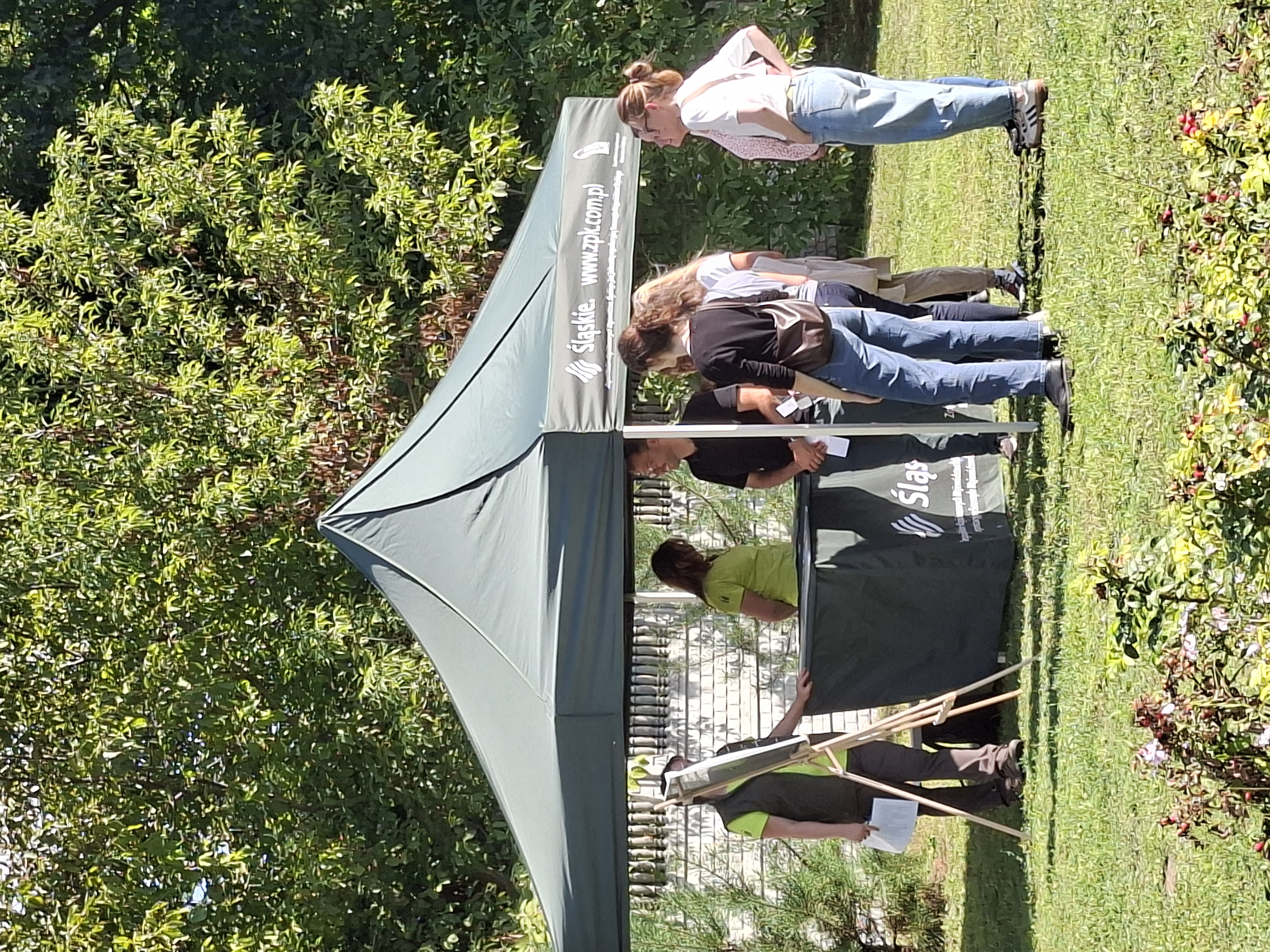 Na zdjęciu widoczny zielony namiot z ustawionym pod nim zielonym stołem. Z lewej strony namiotu na drewnianej sztaludze stoi koło fortuny, przeznaczone do losowania zadań ekologicznych. Przed namiotem stoi pięć osób (kobiet i mężczyzn), które patrzą na koło fortuny i na materiały znajdujące się na stole. Po przeciwnej stronie stołu, pod namiotem, stoją dwie kobiety ubrane w jasnozielone koszulki. W tle duże, zielone drzewo liściaste i betonowe ogrodzenie.