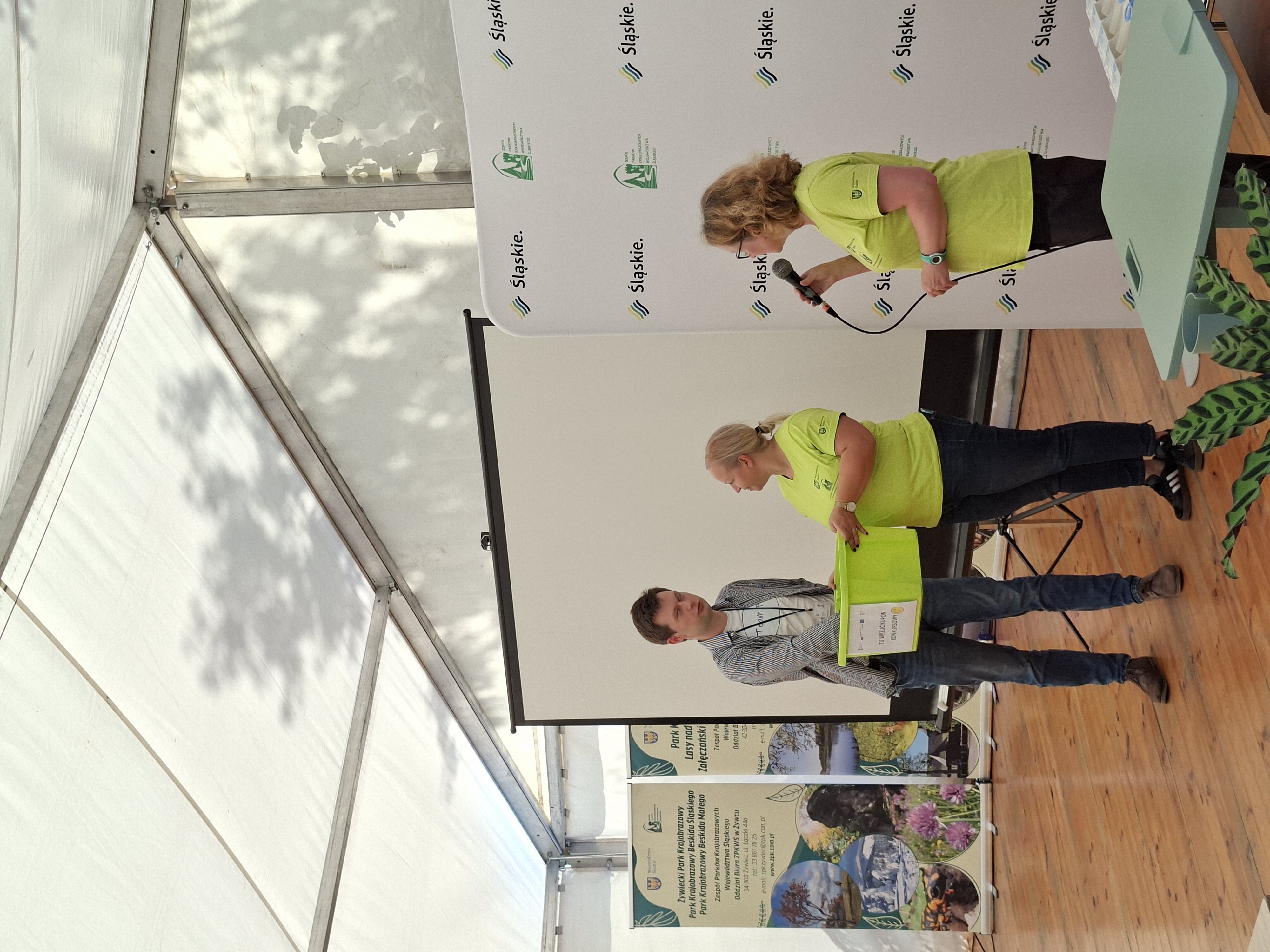 Na zdjęciu widoczne dwie uśmiechnięte kobiety ubrane w jasnozielone koszulki i ciemne spodnie oraz wysoki mężczyzna w jasnej koszulce i niebieskiej koszuli. Wszyscy stoją na tle białego ekranu do wyświetlania prezentacji multimedialnych. Kobieta w okularach, stojąca po prawej stronie trzyma mikrofon w prawej dłoni. Kobieta na środku zdjęcia trzyma jasnozielone, plastikowe pudło, w którym mężczyzna trzyma prawą rękę.