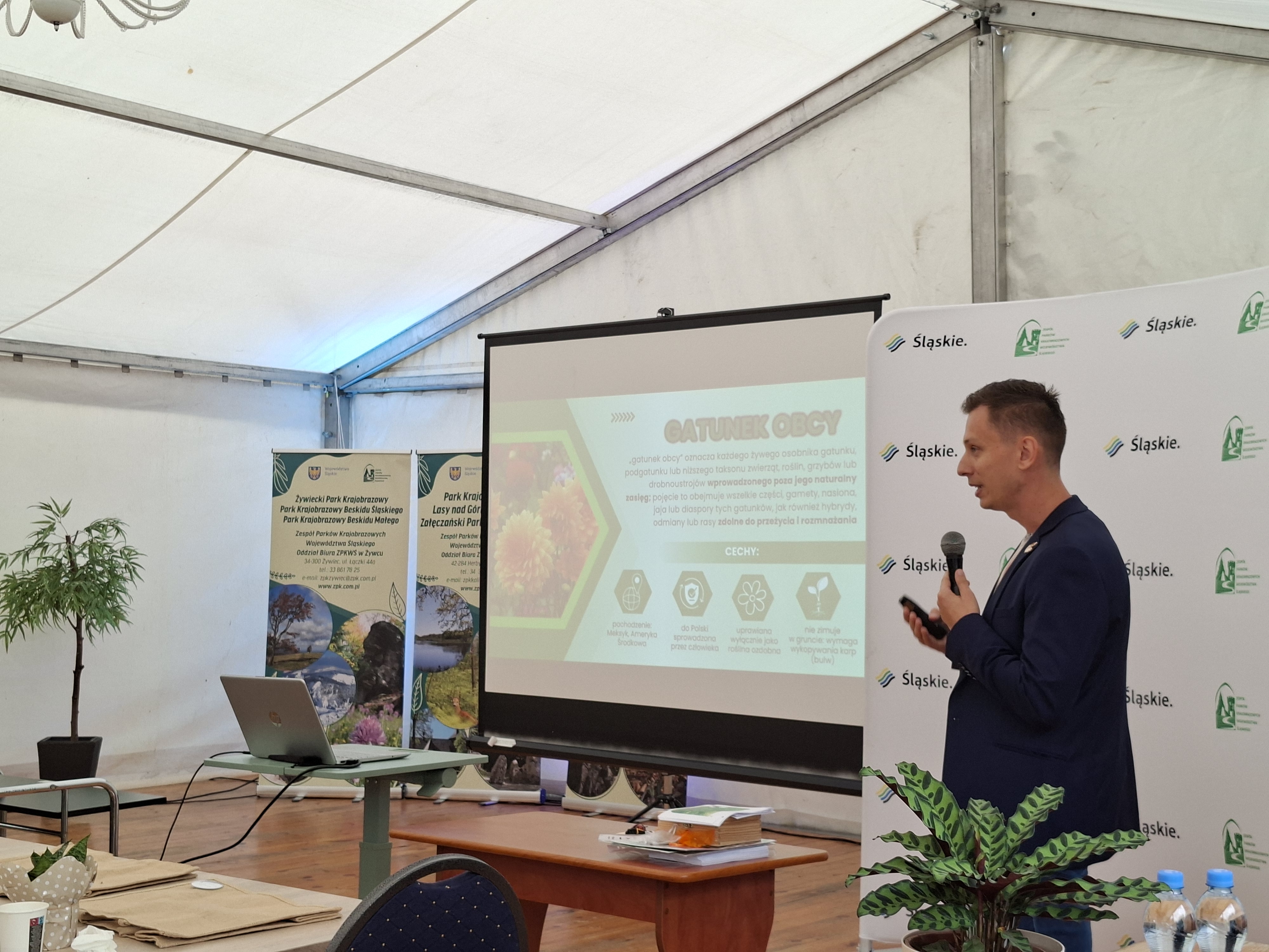 Po prawej stronie stoi mężczyzna z ciemnymi włosami i czarnej marynarce. W dłoni trzyma mikrofon. W środkowej części zdjęcia widoczny ekran z wyświetloną prezentacją multimedialną. W lewej części zdjęcia (na drugim planie) widoczne banery informacyjne o parkach krajobrazowych.