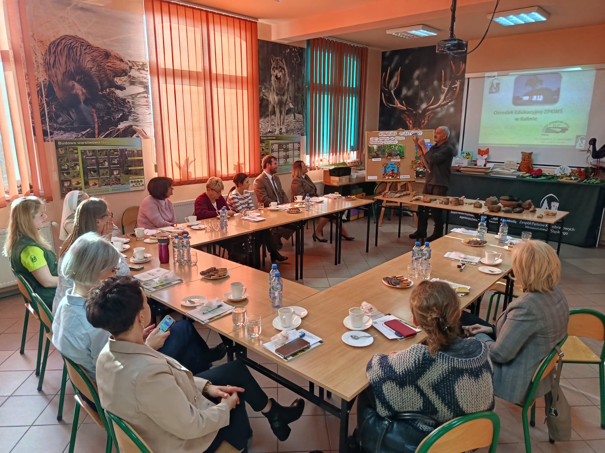 Grupa dorosłych osób siedzących przy kwadratowym stole. na drugim planie pracownik ZPKWŚ przedstawiający ofertę edukacyjną OE ZPKWŚ w Kalinie