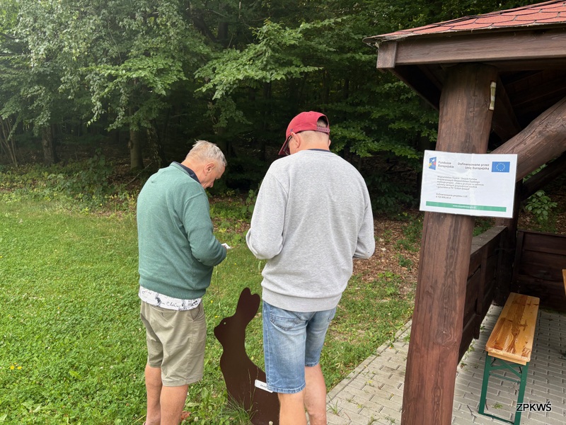 Na zdjęciu widzimy dwóch mężczyzn stojących na świeżym powietrzu, tuż przy skraju lasu. Obaj są ubrani w swobodne, letnie stroje – krótkie spodenki i bluzy. Jeden z nich ma na głowie czerwoną czapkę z daszkiem. Mężczyźni pochylają się nad sylwetką wyciętą z brązowej płyty w kształcie zająca, która stoi na trawie. Wygląda to na element gry terenowej albo edukacyjnej ścieżki przyrodniczej. Obok znajduje się drewniana wiata z ławkami. Do jednego z jej słupów przymocowana jest tablica z logotypami Funduszy Europejskich oraz Unii Europejskiej, informująca o współfinansowaniu projektu.