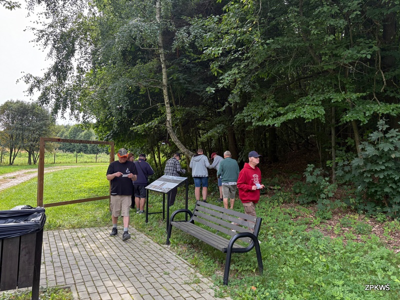Na zdjęciu widać grupę ludzi znajdujących się przy wejściu do lasu. Część osób stoi przy tablicy informacyjnej oraz wchodzi do zalesionego terenu, inni natomiast spacerują w pobliżu. Na pierwszym planie znajduje się ławka oraz kosz na śmieci. Ścieżka z kostki brukowej prowadzi do drewnianej ramy z napisem „PARK KRAJOBRAZOWY”. Po lewej stronie widoczna jest otwarta przestrzeń z trawą i drogą gruntową, a po prawej gęsty las. 