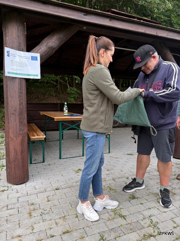 Na zdjęciu widać dwie osoby stojące pod drewnianą wiatą na świeżym powietrzu. Kobieta w zielonej kurtce i dżinsach trzyma worek, z którego coś wyciąga lub do którego coś wkłada mężczyzna ubrany w ciemną bluzę i czapkę z daszkiem. Scena wygląda na element wspólnej zabawy, konkursu albo losowania. W tle, pod wiatą, stoi drewniany stół z ławkami, a na nim butelka napoju oraz kilka przedmiotów. Na jednym z drewnianych słupów widnieje tablica informacyjna z logotypami Funduszy Europejskich i Unii Europejskiej. 