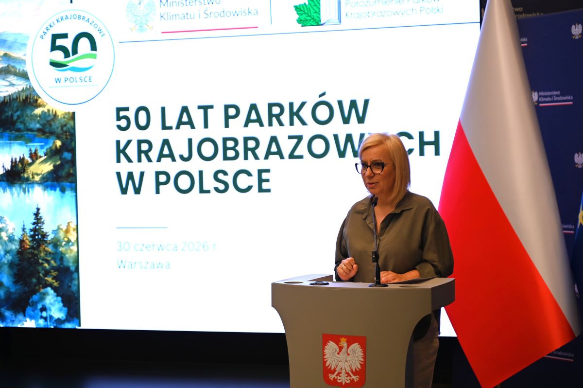 Minister Klimatu i Środowiska Paulina Hennig – Kloska stoi przy mównicy i przemawia do zgromadzonych uczestników konferencji 50 – lecia parków krajobrazowych. W tle napis 50 – lecie parków krajobrazowych.