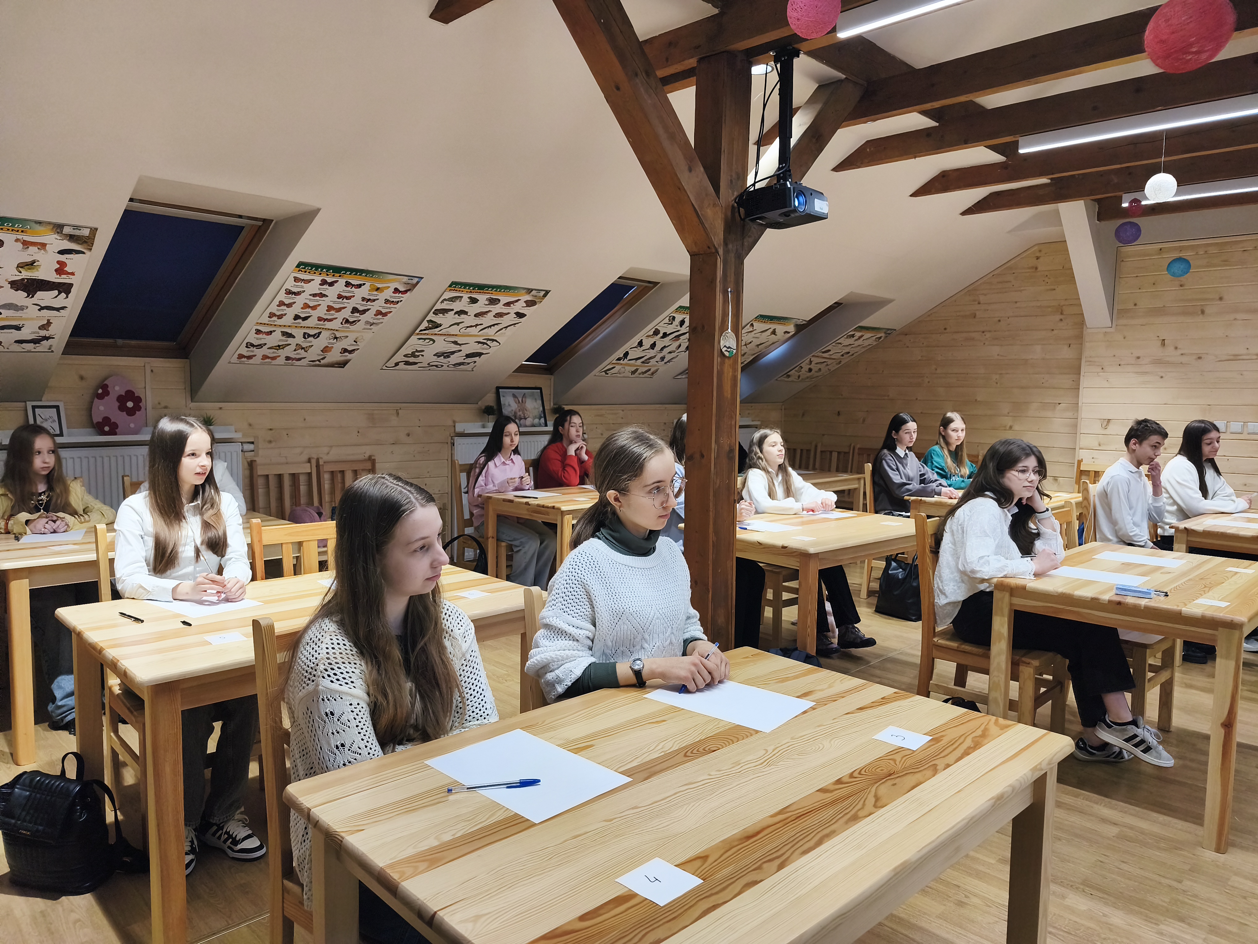 2. W jasnej sali z drewnianymi ścianami i skośnym sufitem kilku uczniów siedzi przy drewnianych ławkach, pracując z kartkami i długopisami. Na suficie wiszą plakaty przedstawiające zwierzęta i owady, a z belki pod sufitem zwisa projektor.
