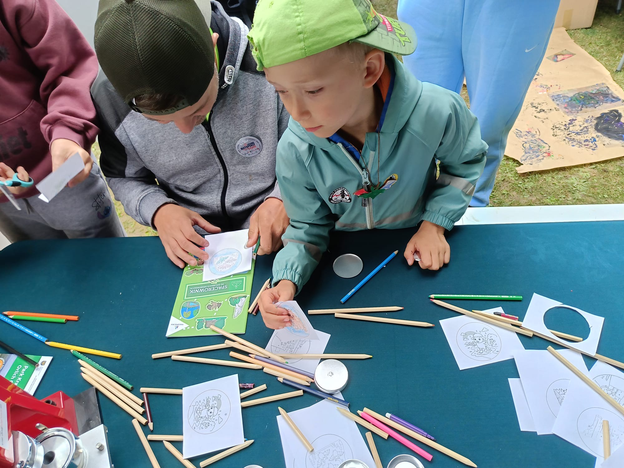 Zdjęcie przedstawia dwójkę chłopców zaangażowanych w zajęcia plastyczne lub warsztaty edukacyjne na świeżym powietrzu. Siedzą przy stole przykrytym zielonym obrusem Na pierwszym planie, po prawej stronie, widać chłopca w jaskrawozielonej czapce z daszkiem oraz niebieskiej, wiatroszczelnej kurtce. Jest pochylony nad białą kartką z wydrukowanym wzorem do kolorowania i używa kredki ołówkowej. Obok niego, po lewej stronie, siedzi starszy chłopak w szarej bluzie z kapturem i ciemnozielonej czapce. Trzyma w ręku zieloną broszurę, na której widać napis. Obaj chłopcy uważnie obserwują czynność wykonywaną przez młodszego. Powierzchnia stołu jest pokryta wieloma przedmiotami: rozrzuconymi kredkami ołówkowymi w różnych kolorach (niebieskie, zielone, różowe, naturalne drewno), małymi, okrągłymi przypinkami przygotowanymi do wykonania lub gotowymi, białą kartką z wydrukowanymi okręgami do kolorowania, a także ulotkami. Z lewej strony zdjęcia widać ramię innej osoby w bordowej bluzie, trzymającej coś w ręku. Z prawej strony, w tle, widoczne są fragmenty nóg innej osoby w jasnoniebieskich spodniach oraz kawałek kartonu leżącego na trawie z kolorowymi rysunkami.
