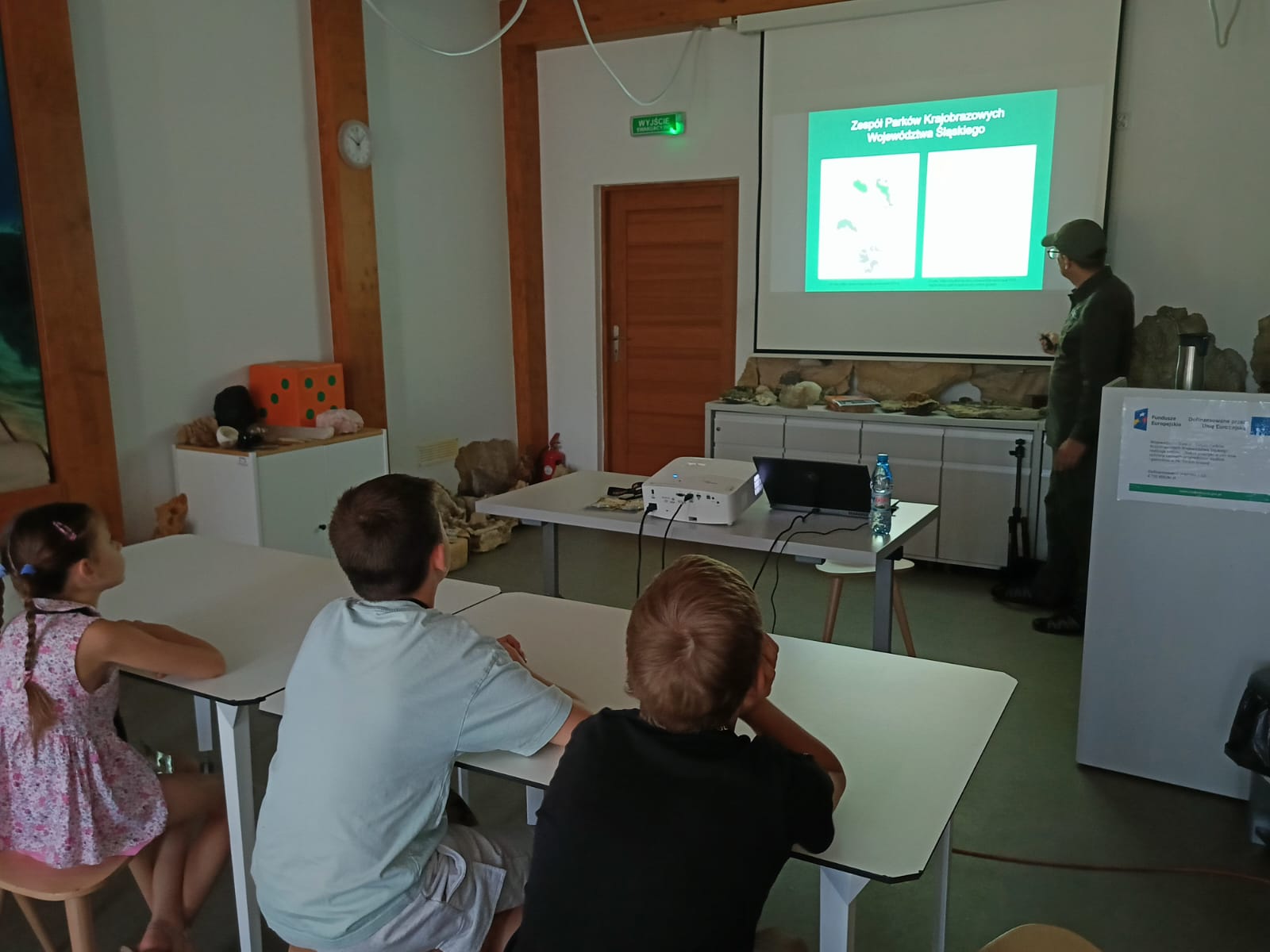 Zdjęcie przedstawia prelekcję edukacyjną w jasnej sali lekcyjnej. W prawym rogu mężczyzna  w zielonym stroju i czapce stoi przy ekranie i wskazuje na wyświetlaną prezentację z napisem „Zespół Parków Krajobrazowych Województwa Śląskiego”. Przed nim, po lewej stronie, tyłem i bokiem do obiektywu siedzą dzieci – uważnie obserwujących pokaz. Na pierwszym planie znajdują się białe stoły szkolne. W tle widać rzutnik, laptopa oraz szafki, na których ułożono okazy skał i minerałów.