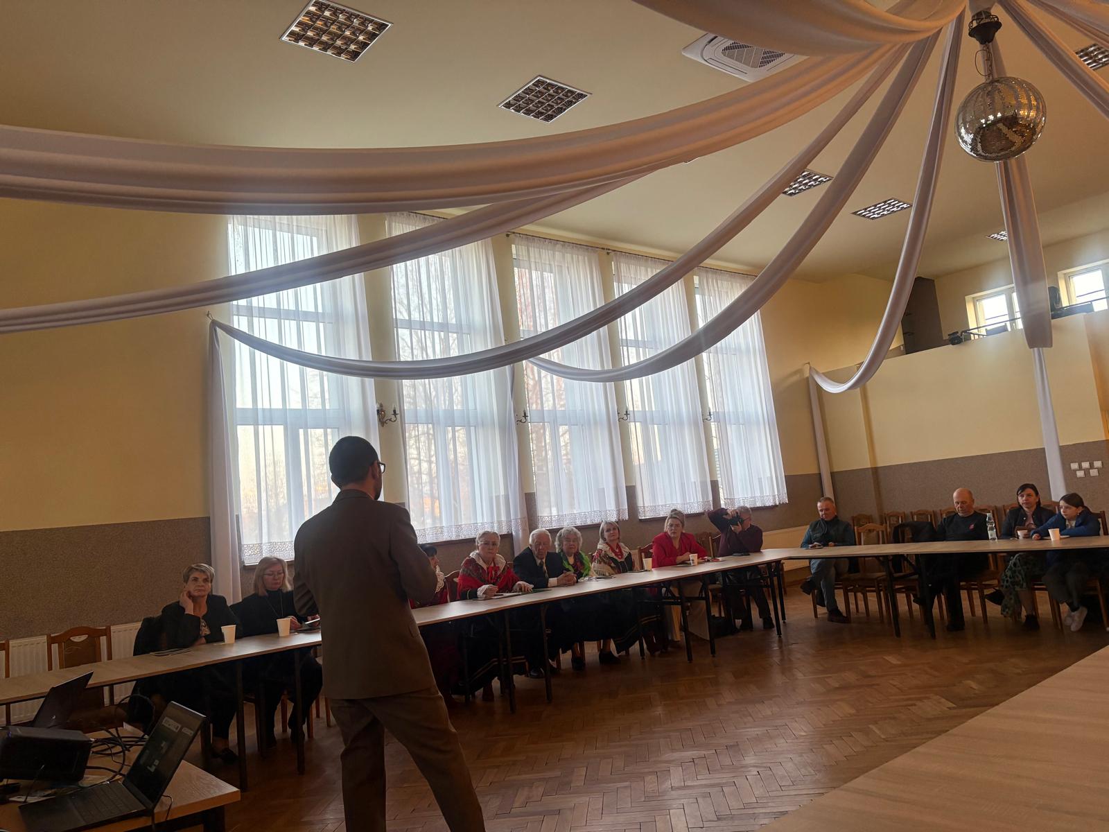 Mężczyzna stojący na środku sali przemawia do grupy osób siedzących przy stołach ustawionych w kształcie podkowy. Spotkanie odbywa się w jasnej sali z dużymi oknami, a z sufitu zwisają dekoracyjne tkaniny i kula dyskotekowa. Na stole obok stoi laptop używany do prezentacji.