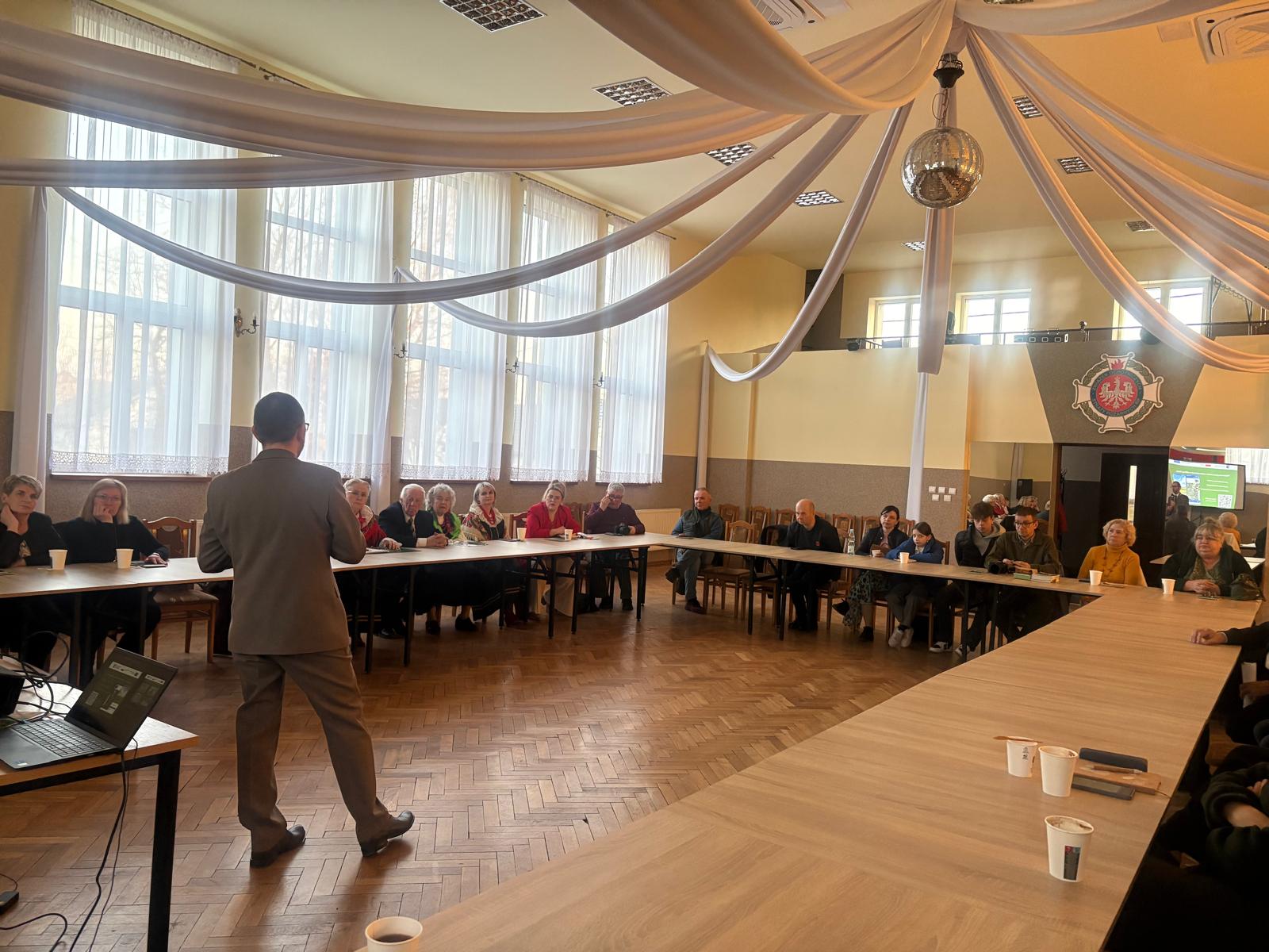 Mężczyzna stojący na środku sali prowadzi spotkanie i przemawia do grupy osób siedzących przy stołach ustawionych w kształcie podkowy. Uczestnicy słuchają prezentacji, a obok znajduje się laptop podłączony do rzutnika. Sala jest przestronna, z dużymi oknami, dekoracyjnymi tkaninami zwisającymi z sufitu i kulą dyskotekową. W tle widoczny jest herb na ścianie oraz ekran z wyświetlaną prezentacją.