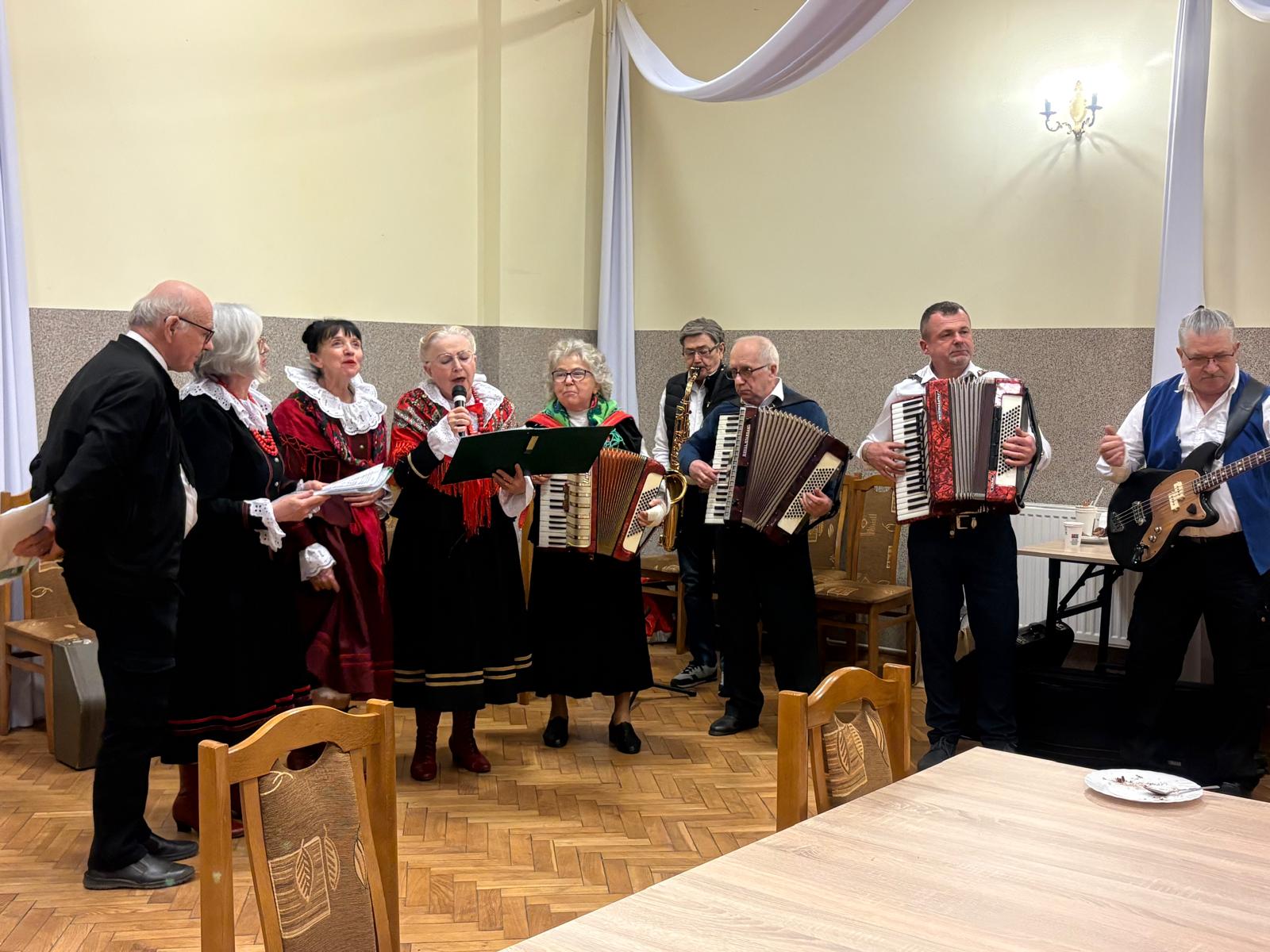 Grupa starszych osób występuje w sali, śpiewając i grając muzykę. Kobiety w tradycyjnych strojach ludowych śpiewają do mikrofonu, a obok mężczyźni grają na akordeonach, saksofonie i gitarze basowej.