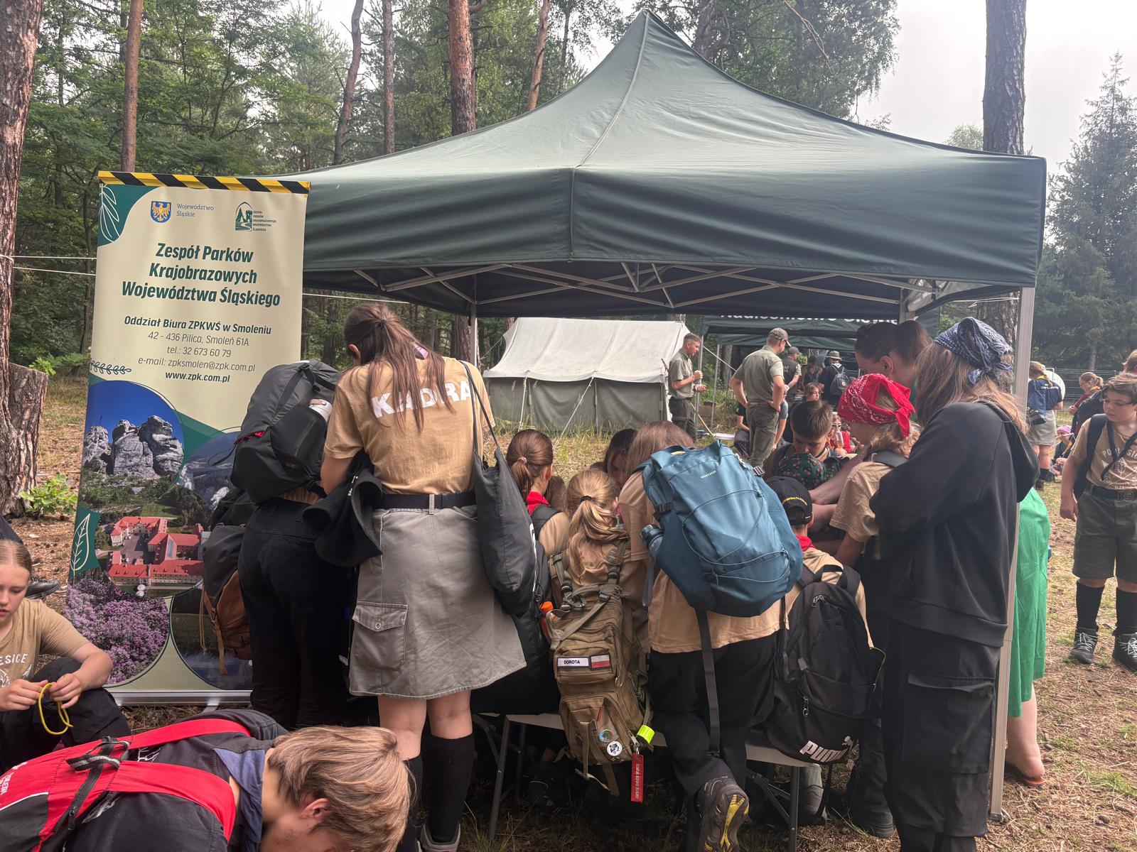 Stoisko edukacyjne Zespołu Parków Krajobrazowych Województwa Śląskiego (ZPKWŚ) podczas wydarzenia w leśnym obozowisku. Liczna grupa harcerzy w beżowych koszulkach, mundurach i z plecakami stoi tyłem i bokiem do obiektywu, tłocząc się pod zielonym namiotem promocyjnym. Po lewej stronie stoi pionowy baner informacyjny z logo Województwa Śląskiego, nazwą instytucji oraz danymi kontaktowymi Oddziału Biura ZPKWŚ w Smoleniu. Jedna z harcerek stojących tyłem ma na koszulce wyraźny napis „KADRA”. W tle, na leśnej polanie, widoczne są duże szare namioty wojskowe oraz inni uczestnicy i instruktorzy w mundurach.