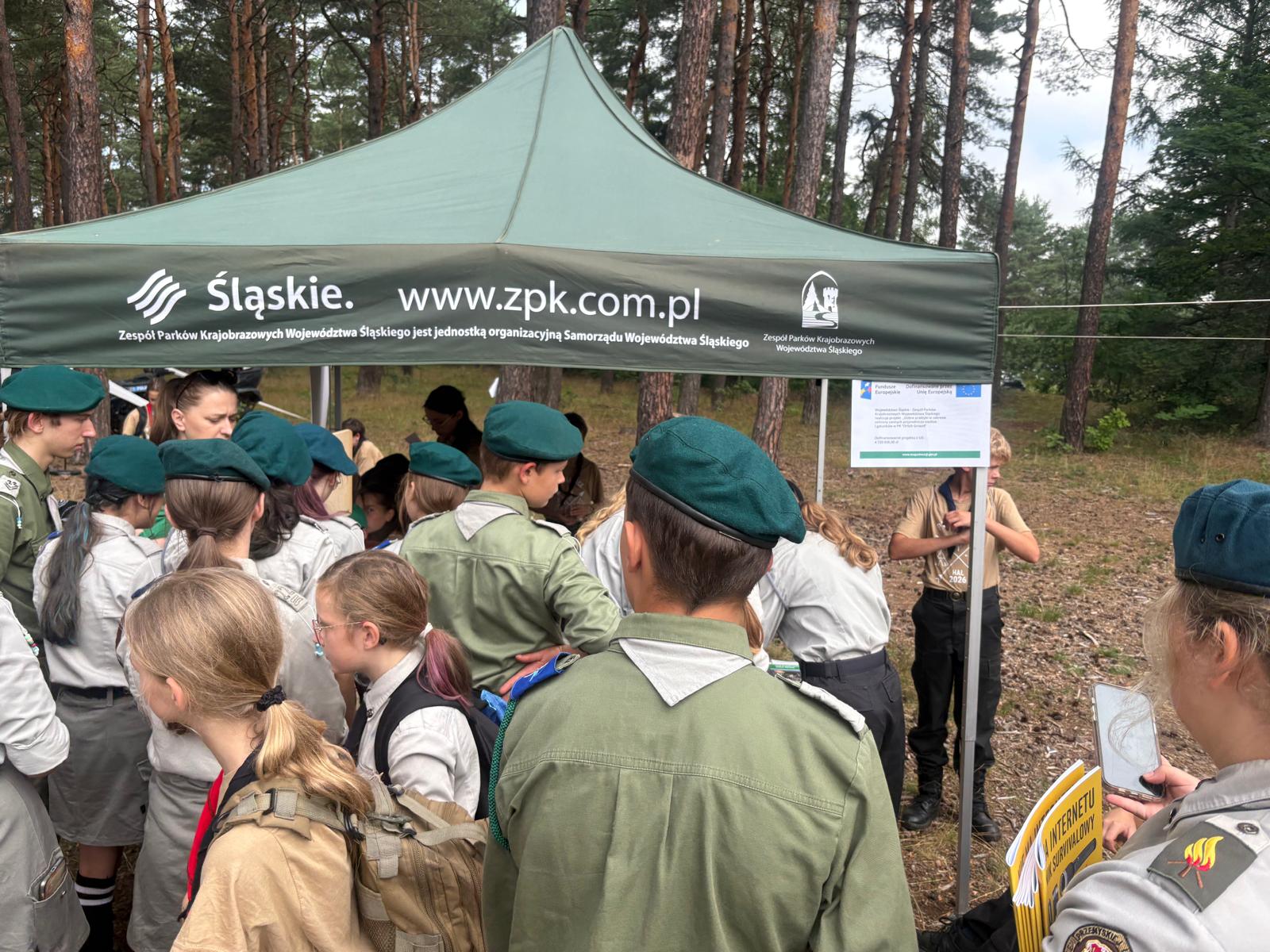 Stoisko edukacyjne Zespołu Parków Krajobrazowych Województwa Śląskiego (ZPKWŚ) w leśnym otoczeniu. Liczna grupa harcerzy, w większości ubranych w oficjalne, szaro-zielone mundury oraz ciemnozielone berety, stoi pod zielonym namiotem promocyjnym instytucji. Na prawym słupku stelaża namiotu zawieszony jest biały plakat informacyjny o dofinansowaniu projektu przez Unię Europejską (Fundusze Europejskie). Po prawej stronie, na pierwszym planie, widoczna jest harcerka trzymająca w dłoniach telefon komórkowy oraz żółtą broszurę informacyjną z widocznym napisem zawierającym słowo „SURVIVALOWY”. Tło zdjęcia stanowią gęsto rosnące sosny.