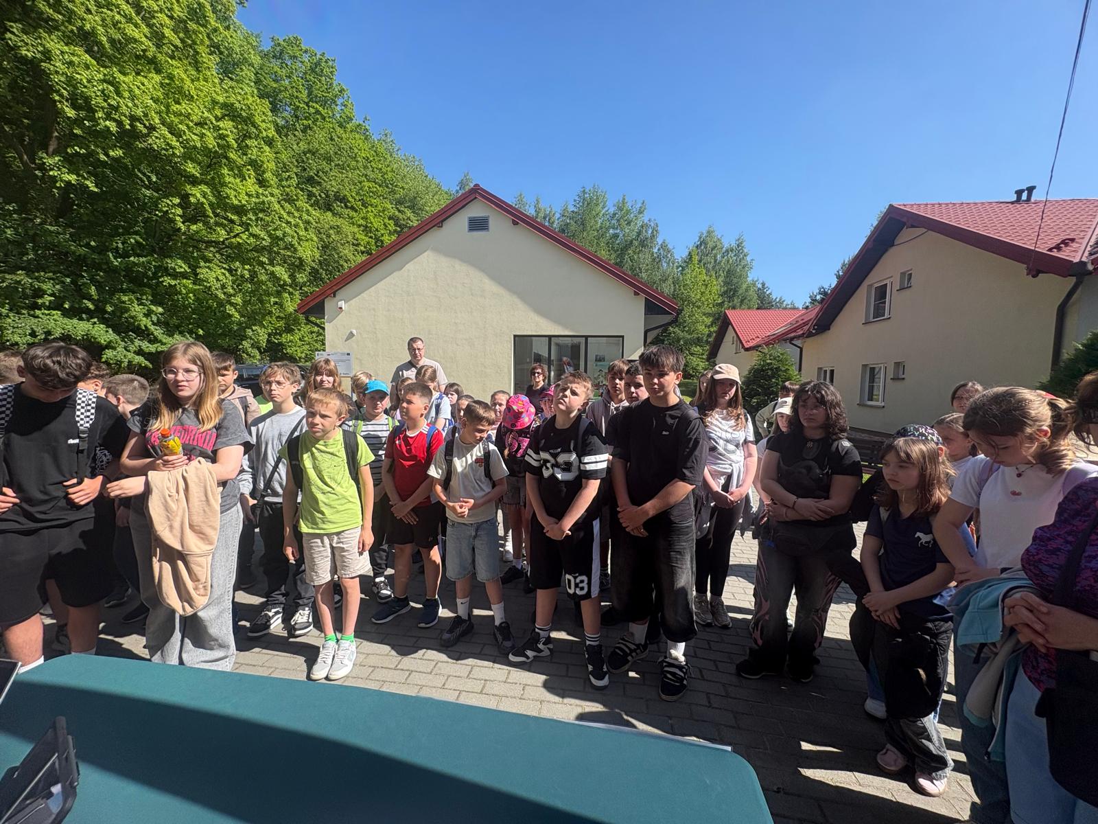 Zdjhęcie przedstawia dużą grupę dzieci zgromadzona na wybrukowanym placu przez budynkami ZPKWŚ Oddział w Smoleniu z jasnymi elewacjami i spadzistymi dachami koloru czerwonego. Dzieci, obrani w swobodne, letnie stroje sportowe i plecaki, stoja w skupieniu i patrzą w jednym kierunku, słuchając zasad gry terenowej przedstawianej przez organizatorów. Przed dziećmi stoi stół w kolorze zielonym. Cała scena rozgrywa się w pełnym słońcu, pod bezchmurnym niebiem, a w drugim planie widać gęste, zielone drzewa.