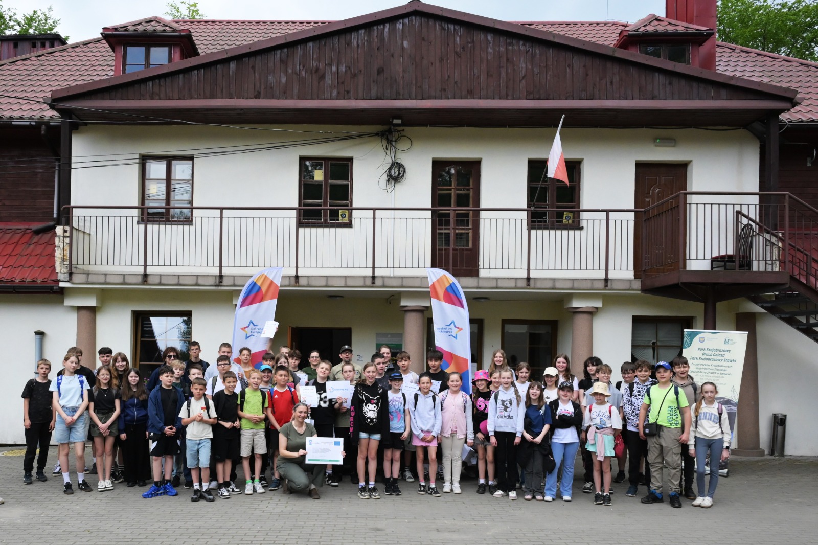 Na zdjęciu widoczna jest liczna grupa dzieci wraz z opiekunamiy, ustawiona przed budynkiem głównym ZPKWŚ Oddział w Smoleniu. Uczestnicy pozują do współnego zdjęcia pamiątkowego, trzymając dyplomu i materiały związane z wydarzenim Funduszowy Maj - gra terenowa. Przed wejściem ustawione sa flagi promocyjne oraz tablica informacyjna. Fotografia przedstawiam zakońcenie gry terenowej.