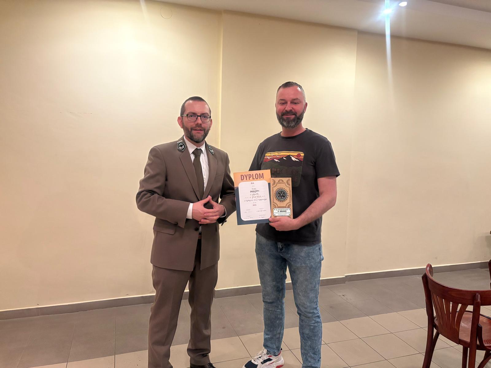 Dwóch mężczyzn stoi w jasnej sali o kremowych ścianach. Mężczyzna po lewej stronie ma na sobie elegancki zielony mundur z krawatem i okularami. Mężczyzna po prawej jest ubrany bardziej swobodnie w czarny T-shirt z kolorową grafiką, dżinsy i sportowe buty. Trzyma w rękach dyplom oraz ozdobną statuetkę. Obaj stoją obok siebie i patrzą w stronę aparatu. 