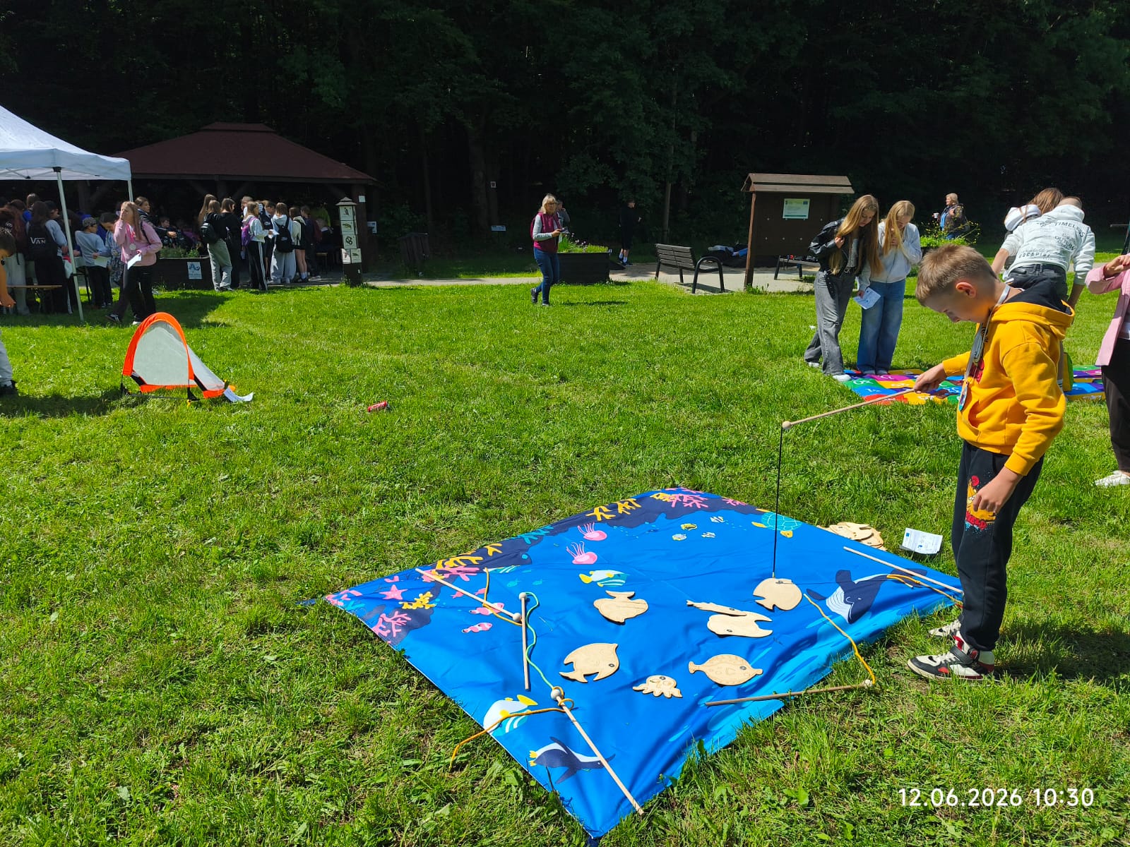 Uczestnicy plenerowego pikniku biorą udział w grach zręcznościowych na polanie przy oddziale ZPKWŚ w Smoleniu. Na pierwszym planie po prawej stronie chłopiec w żółtej bluzie bawi się w łowienie rybek, używając drewnianej wędki i starając się podnieść drewniane rybki rozłożone na dużej, niebieskiej macie animacyjnej imitującej wodę. W tle po prawej stronie dziewczęta korzystają z innej kolorowej maty do gier. Dalej, w głębi polany pod lasem, widoczne są drewniane altany, namiot oraz zgromadzone grupy dzieci i młodzieży.
