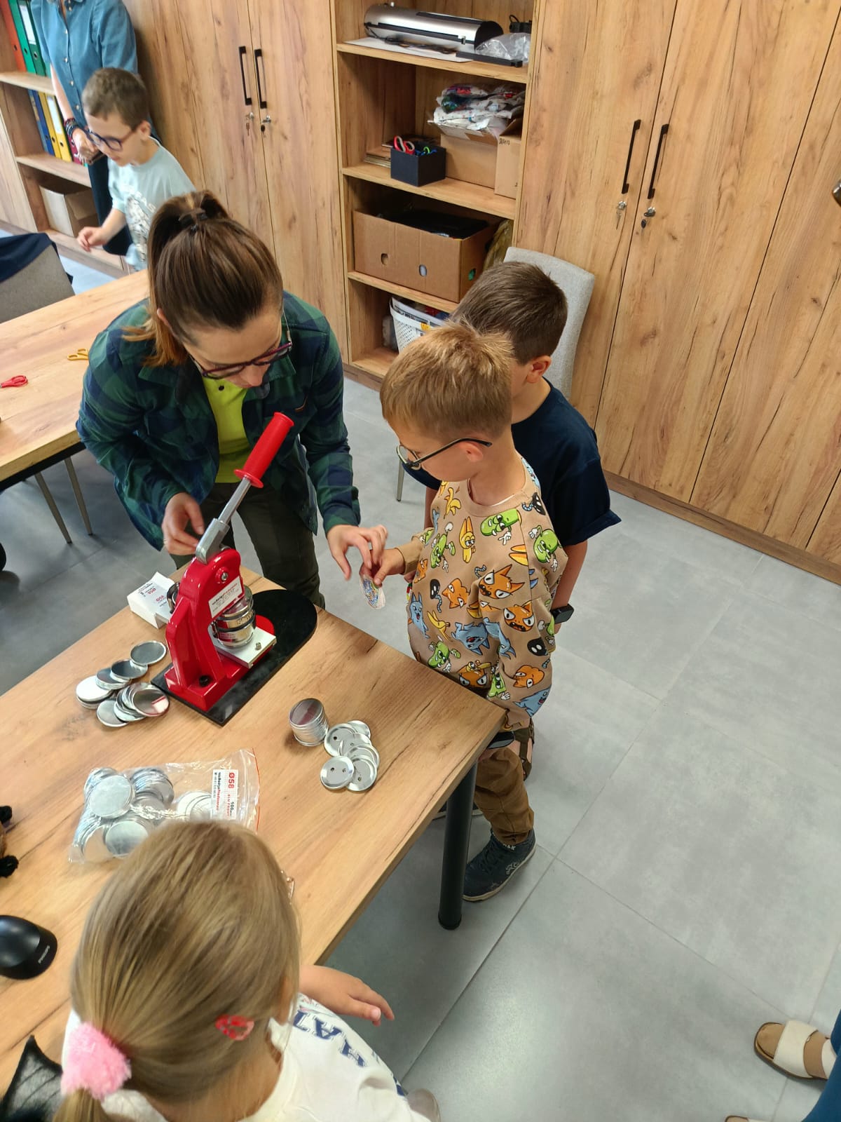 Zdjęcie przedstawia warsztaty edukacyjno-manualne dla dzieci, odbywające się w jasnym pomieszczeniu. Dorosła kobieta w okularach, zielonym t-shircie i rozpiętej zielono-granatowej koszuli w kratę stoi przy drewnianym stole. Pochyla się w stronę dwóch stojących obok chłopców i pokazuje jednemu z nich, jak przygotować element do maszyny. Na stole znajduje się profesjonalna, czerwona prasa (maszynka) do robienia przypinek z długą, czerwoną rączką. Obok niej leżą ułożone w stosiki oraz rozsypane metalowe komponenty do przypinek (spody i kapsle) oraz woreczek foliowy z kolejnymi elementami. Chłopiec na pierwszym planie, w okularach i beżowej bluzie we wzory z rysunkowymi postaciami, przygląda się trzymanemu w dłoni krążkowi. Tuż za nim stoi drugi chłopiec w ciemnej koszulce. W dolnej części kadru widoczna jest głowa i ramię dziewczynki z jasnymi włosami spiętymi różową gumką, która również obserwuje proces. W tle widać drewniane meble biurowe (regał z segregatorami i pudełkami, szafę) oraz fragmenty innych osób uczestniczących w spotkaniu. Podłoga w pomieszczeniu wyłożona jest szarymi płytkami.