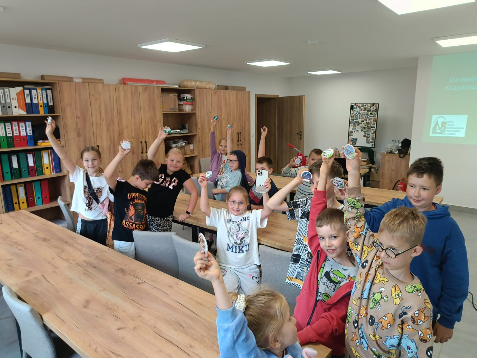Zdjęcie przedstawia grupę kilkanaściorga dzieci w wieku szkolnym pozujących w sali warsztatowej. Dzieci stoją za podłużnymi, drewnianymi stołami i patrzą w stronę obiektywu z uśmiechem, prezentując do zdjęcia wykonane przez siebie okrągłe przypinki z różnymi wzorami (m.in. rysunkami i motywami przyrodniczymi), które trzymają w uniesionych dłoniach. Jedna z dziewczynek w centrum trzyma również telefon komórkowy skierowany aparatem w stronę fotografa. Na pierwszym planie po prawej stronie widać chłopców z bliska – jeden w okularach i wzorzystej beżowej bluzie, drugi w czerwonej rozpinanej bluzie z kapturem, a za nimi kolejny chłopiec w niebieskiej bluzie. Po lewej stronie, w głębi sali, dzieci stoją w rzędzie wzdłuż stołów. W tle jasnego pokoju znajdują się wysokie, drewniane szafy i regały biurowe wypełnione kolorowymi segregatorami oraz pudełkami. Po prawej stronie na ścianie widoczny jest fragment wyświetlanego z rzutnika slajdu z widocznym napisem „Działalność...” oraz logo Zespołu Parków Krajobrazowych Województwa Śląskiego. Pomieszczenie oświetlone jest sufitowymi lampami kasetonowymi.
