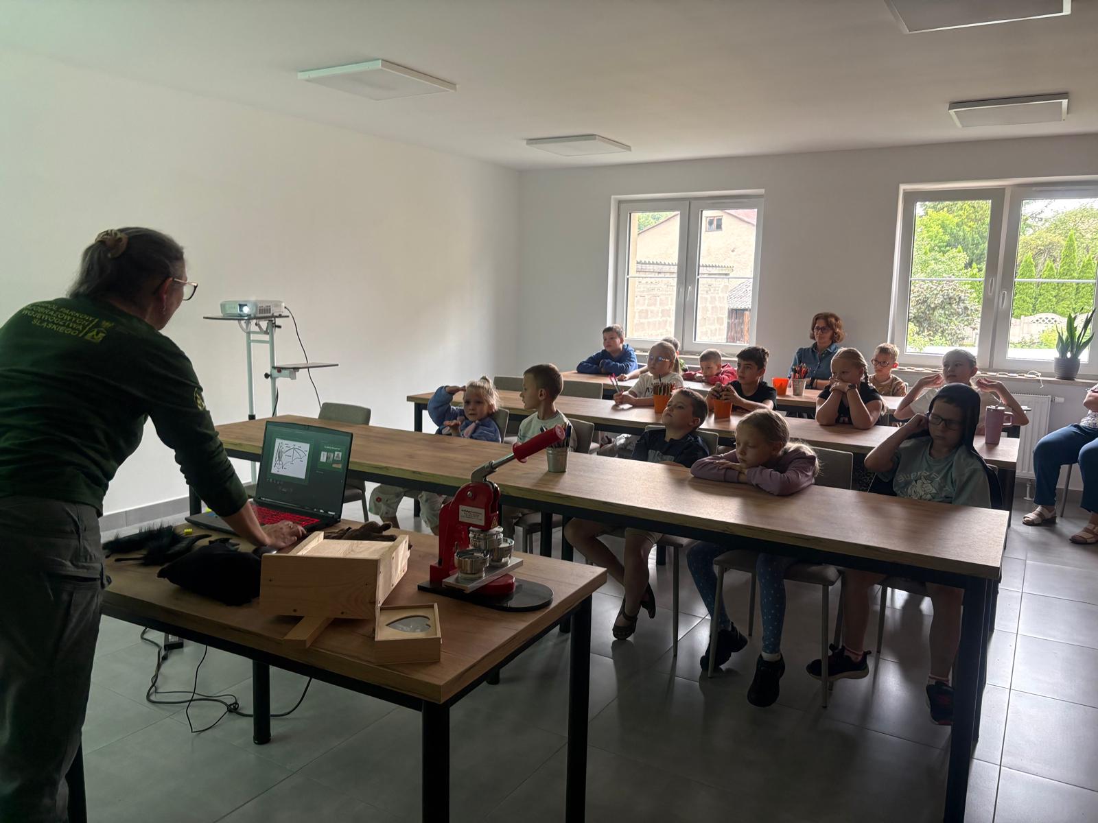 Zdjęcie przedstawia prelekcję edukacyjną dla grupy dzieci w jasnej sali szkoleniowej. Po lewej stronie, tyłem do obiektywu, stoi pracownica Zespołu Parków Krajobrazowych Województwa Śląskiego ubrana w ciemnozieloną koszulkę z logotypem instytucji na plecach. Kobieta obsługuje otwartego laptopa stojącego na biurku, na którym widać slajd prezentacji. Na biurku prowadzącej, obok laptopa, znajdują się pomoce dydaktyczne: czerwona maszynka do robienia przypinek, drewniane pudełka, w tym jedno z wieczkiem w kształcie serca, oraz czarne rekwizyty (prawdopodobnie sztuczne futerko). W głębi, na podwyższeniu, stoi rzutnik multimedialny. W rzędach drewnianych stołów siedzi kilkanaścioro dzieci w wieku szkolnym oraz dwie dorosłe kobiety. Dzieci siedzą przodem do prowadzącej, z uwagą słuchając wykładu i patrząc przed siebie. Na stołach przed niektórymi dziećmi stoją plastikowe kubki z przyborami do pisania. Sala jest dobrze oświetlona naturalnym światłem wpadającym przez dwa duże, białe okna po prawej stronie. Pomieszczenie ma białe ściany i szarą, kaflową podłogę.