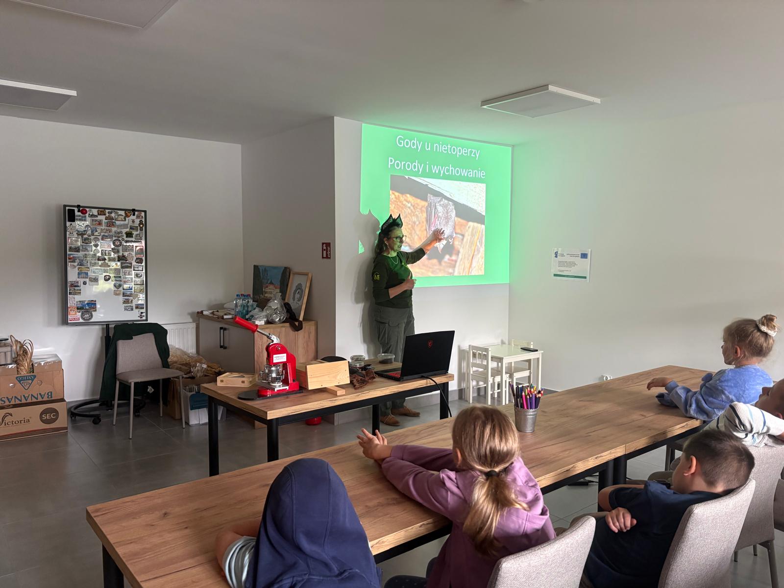 Zdjęcie przedstawia prelekcję edukacyjną dla dzieci prowadzoną w jasnej sali warsztatowej. W centrum kadru stoi pracownica Zespołu Parków Krajobrazowych Województwa Śląskiego (ZPKWŚ) ubrana w ciemnozielony komplet mundurowy. Na głowie ma założoną opaskę imitującą uszy nietoperza. Kobieta wskazuje ręką na wyświetlany z rzutnika na ścianie slajd prezentacji o zielonym tle z widocznym napisem „Gody u nietoperzy. Porody i wychowanie” oraz zdjęciem przedstawiającym wiszącego nietoperza. Przed prowadzącą stoi drewniane biurko z otwartym laptopem, czerwoną maszynką do robienia przypinek, drewnianym pudełkiem oraz innymi rekwizytami. Na pierwszym planie, tyłem do obiektywu, siedzi przy długich, drewnianych stołach kilkoro dzieci, które z uwagą oglądają prezentację. Na stole stoi metalowy kubek z kolorowymi kredkami. W tle sali widoczna jest tablica magnetyczna z przypiętymi magnesami i pamiątkami, mały biały stolik z krzesełkami dla dzieci oraz zawieszony na ścianie biały plakat informacyjny Funduszy Europejskich.