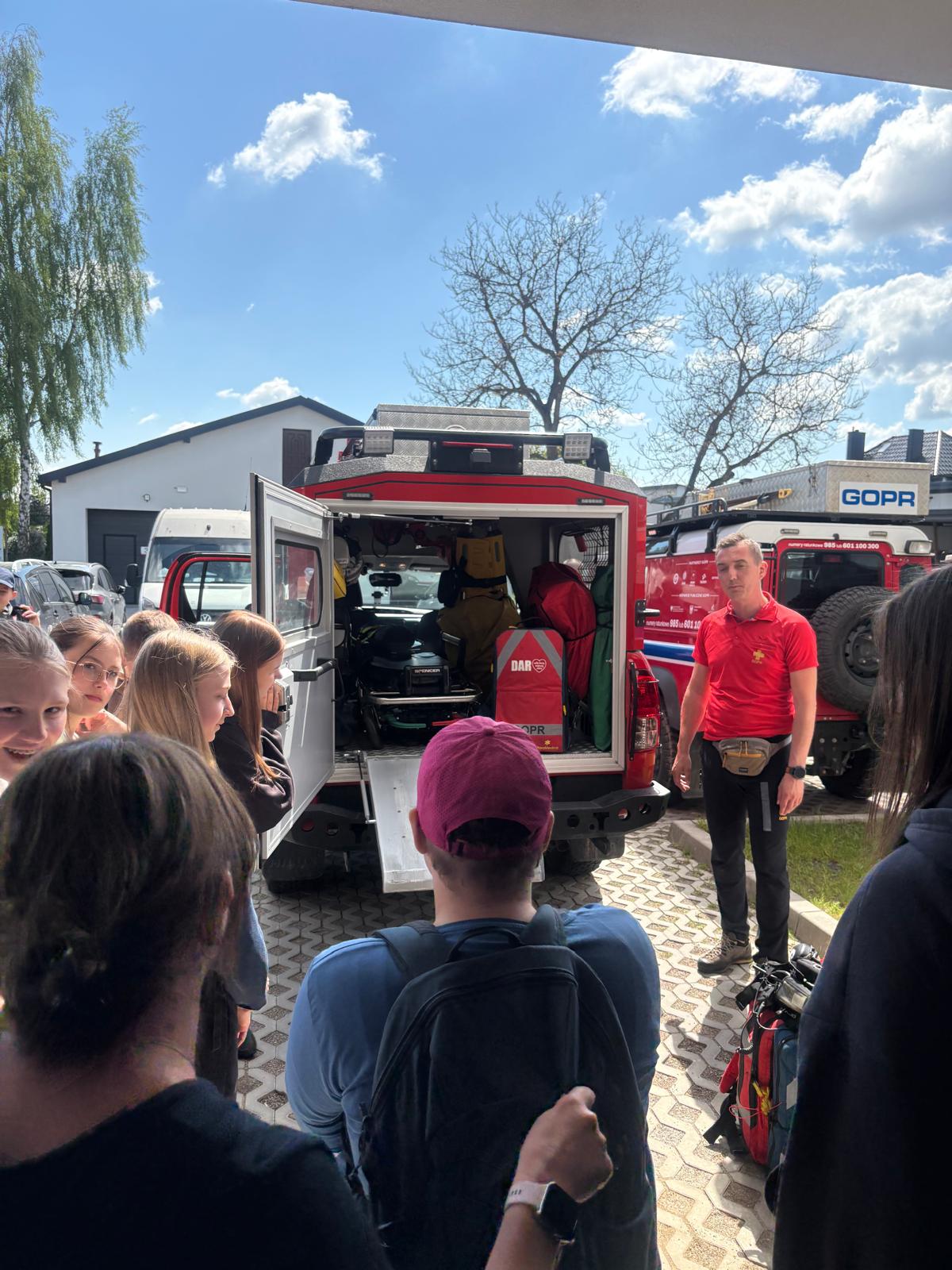 Zdjęcie przedstawia prezentację specjalistycznego sprzętu ratowniczego dla grupy dzieci i młodzieży na świeżym powietrzu w słoneczny dzień. Głównym elementem jest otwarty tył czerwonego samochodu terenowego GOPR, w którym widoczne jest wyposażenie medyczne i ratunkowe, w tym nosze, torby i liny. Obok pojazdu stoi ratownik górski w czerwonej koszulce polo i czarnych spodniach, który opowiada o pracy i sprzęcie. Zgromadzona wokół młodzież z zaciekawieniem zagląda do wnętrza samochodu. W tle widać drugi pojazd ratowniczy z napisem „GOPR”, budynki oraz drzewa pod błękitnym niebem.