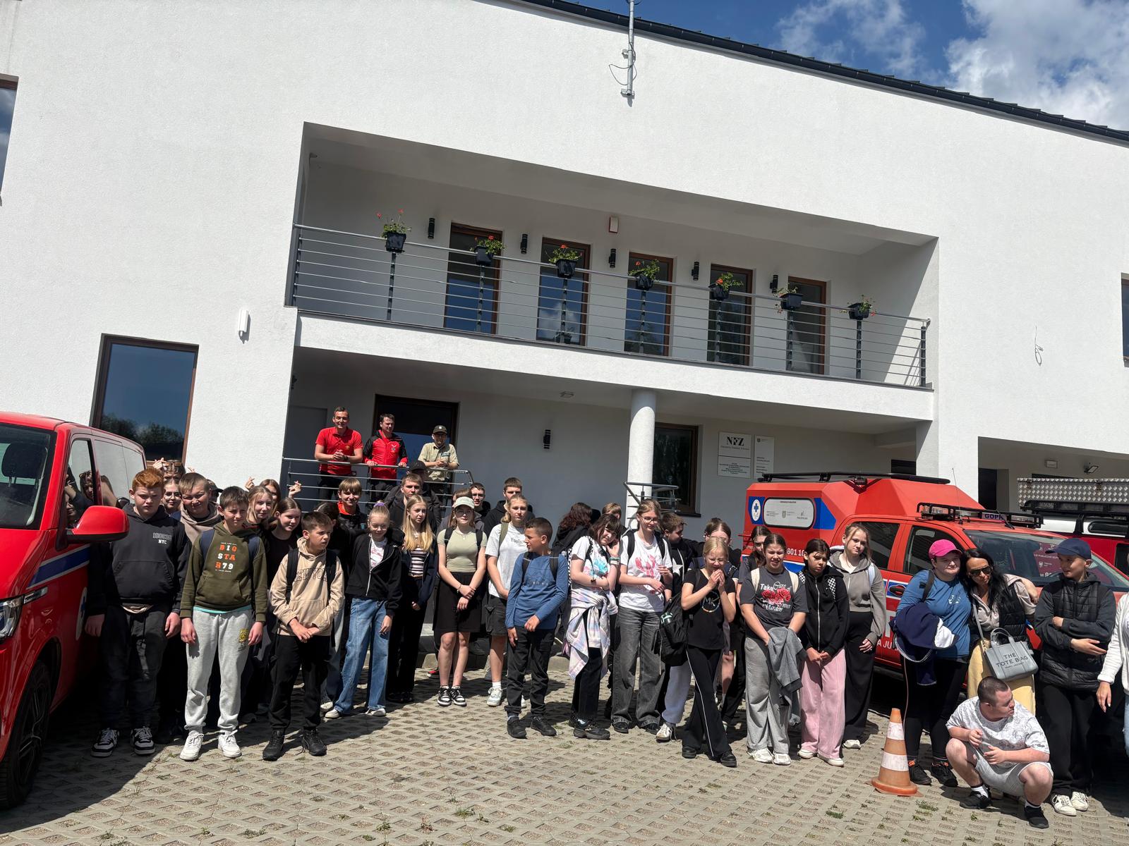 Zdjęcie przedstawia dużą grupę dzieci, młodzieży oraz dorosłych pozujących do wspólnej fotografii pamiątkowej na wybrukowanym placu przed nowoczesnym, białym budynkiem z balkonem. Uczestnicy spotkania stoją ustawieni w kilku rzędach, a po obu stronach grupy widoczne są czerwone, specjalistyczne samochody ratownicze. W tle, na schodach prowadzących do budynku, stoi trzech mężczyzn, w tym ratownicy w charakterystycznych czerwonych strojach. Całość sceny rozgrywa się w słoneczny dzień pod błękitnym niebem z białymi chmurami.
