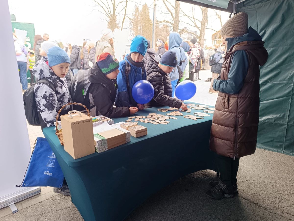 Zdjęcie przedstawia scenę na świeżym powietrzu podczas wydarzenia plenerowego, jest chłodny dzień (uczestnicy są ubrani w zimowe kurtki, czapki i szaliki). Na pierwszym planie znajduje się stoisko przykryte zielonym obrusem, ustawione pod namiotem. Przy stole stoi grupa kilku dzieci w wieku szkolnym. Mają na sobie zimowe ubrania: puchowe kurtki, czapki i plecaki. Troje z nich trzyma niebieskie balony. Dzieci pochylają się nad stołem i oglądają lub układają drewniane elementy – małe, płaskie klocki i większe wycięte kształty, przypominające puzzle lub edukacyjną układankę. Po prawej stronie stołu stoi dorosła osoba (prawdopodobnie prowadząca stoisko), ubrana w długą, brązową kamizelkę, zieloną kurtkę i czapkę. Osoba ta obserwuje dzieci i jest zwrócona w ich stronę. Na stole, oprócz drewnianych elementów, znajdują się również inne przedmioty: małe pudełka, stosy ulotek lub kart oraz koszyk. Z przodu stołu stoi większe drewniane pudełko z uchwytem.