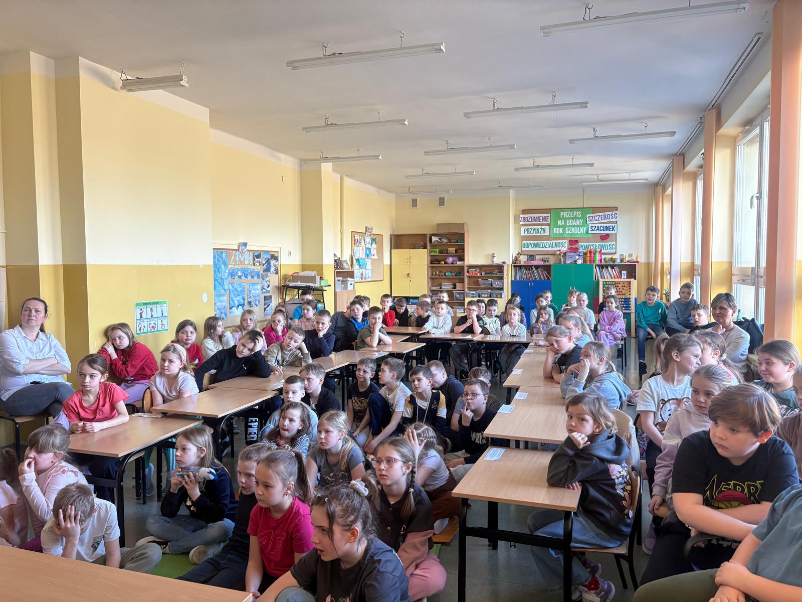 Grupa dzieci siedzi w szkolnej klasie przy ławkach i na podłodze, uważnie słuchając zajęć. W sali znajdują się szkolne pomoce dydaktyczne, półki z książkami oraz kolorowe plakaty edukacyjne na ścianach. Uczniowie patrzą w stronę prowadzącego zajęcia.