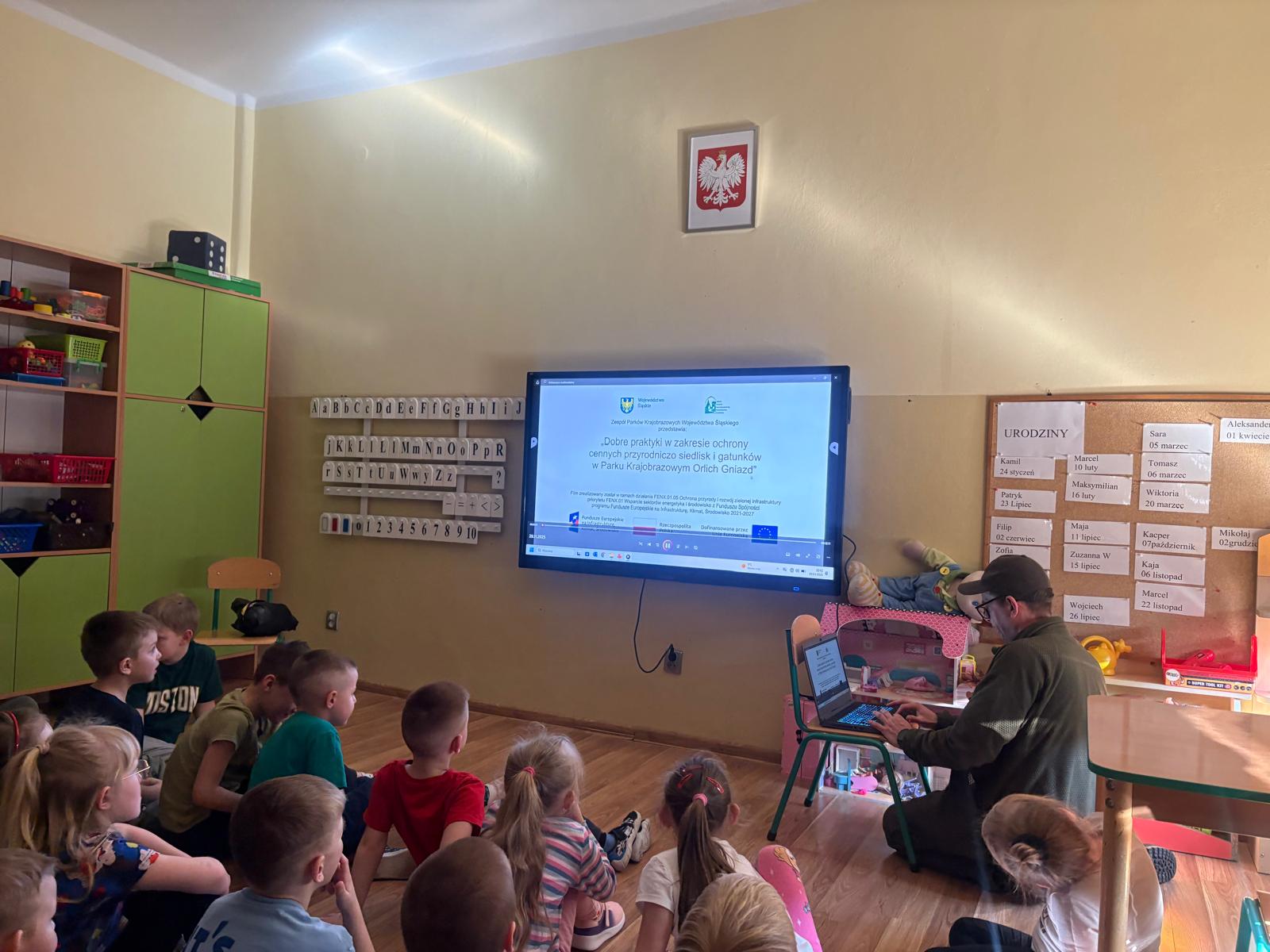 Na zdjęciu widać salę przedszkolną lub wczesnoszkolną podczas zajęć edukacyjnych.  Na podłodze siedzi grupa małych dzieci, które uważnie patrzą na duży ekran zawieszony na ścianie. Na ekranie wyświetlana jest film. Przed ekranem siedzi mężczyzna przy laptopie, który włącza film. 